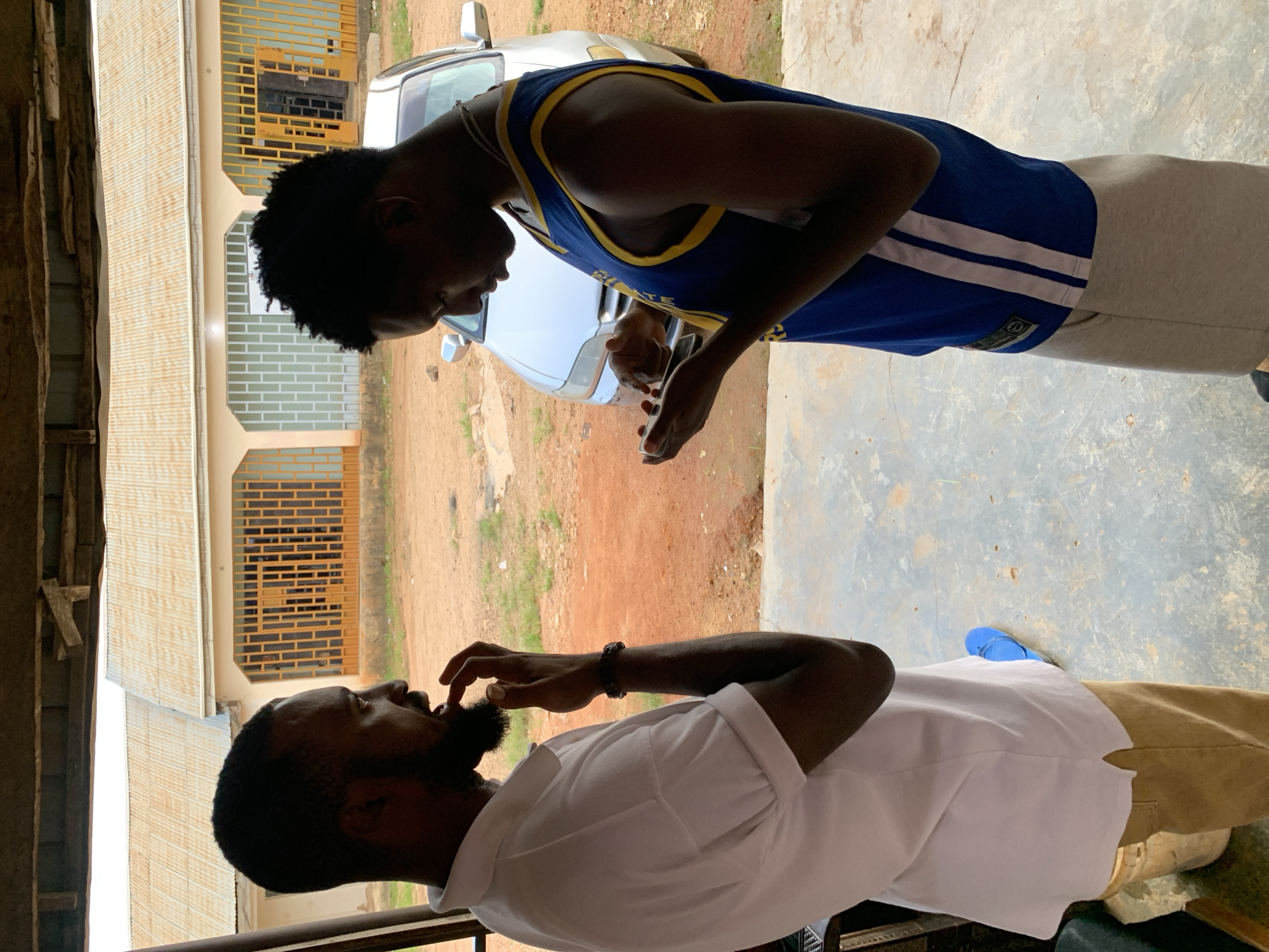 Two young men are standing in an outdoor, covered area, possibly a veranda or carport. The subject in the foreground, wearing a white short-sleeved collared shirt and khaki pants, has a beard and is looking up, with his right hand near his mouth. He is wearing a beaded bracelet on his right wrist and a blue sandal on his left foot.
The second subject, standing on the left and looking down at the first, is wearing a blue and yellow basketball jersey and light-colored shorts. His left hand is extended towards the first man, and he appears to be holding a smartphone in his right hand.
Behind them, a silver car is visible, parked on a dirt and gravel ground with sparse vegetation. A building with corrugated metal roofing and light-colored walls with barred windows is also in the background. The scene appears to be in Jos, Nigeria. The lighting suggests it is daytime. There is no visible text in the image.