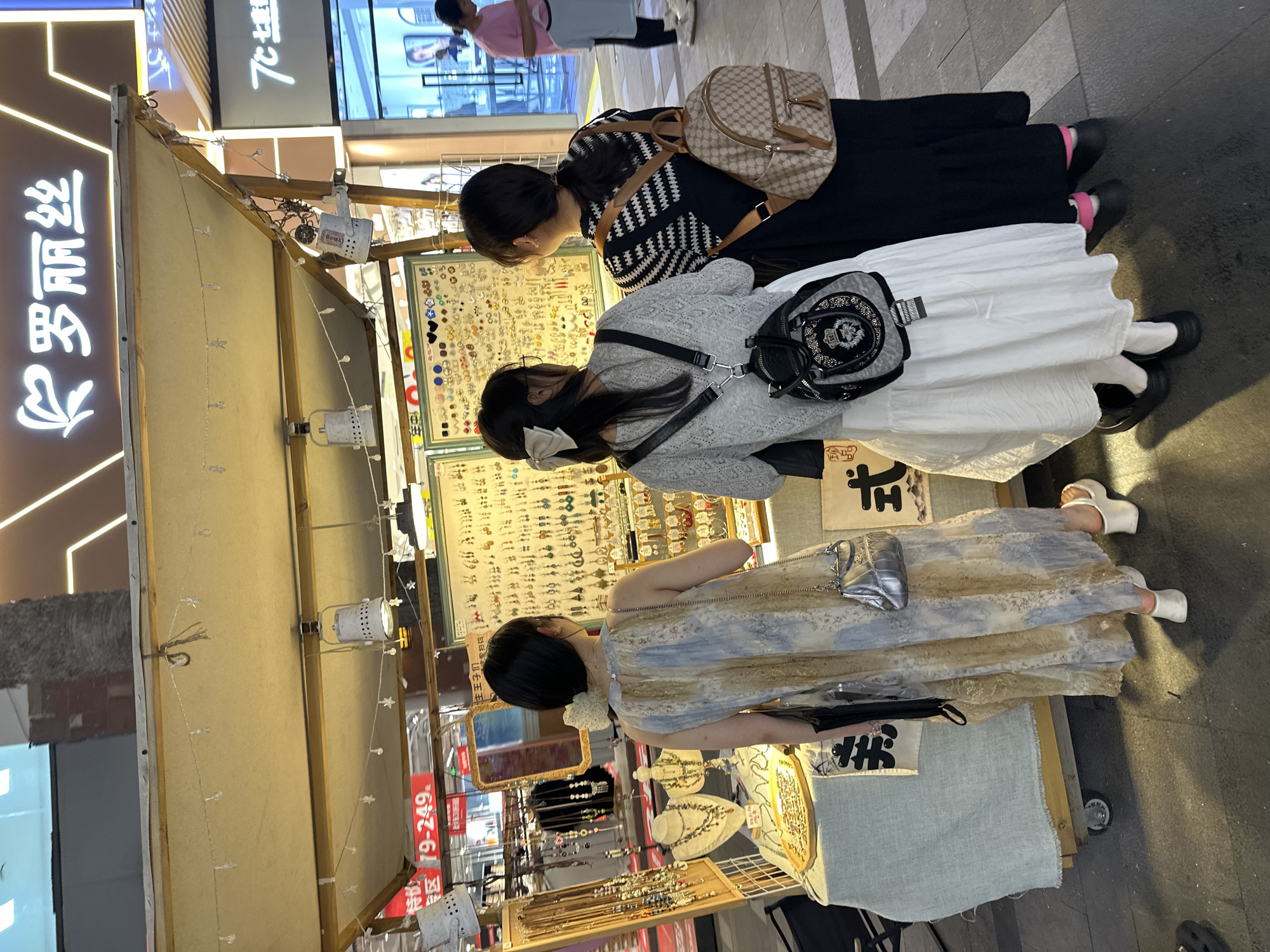 In the Chongqing High-tech Zone, China, a street vendor displays an assortment of jewelry. Three young women, viewed from behind, are inspecting the merchandise at a brightly lit stall. The woman on the left wears a flowing, abstract-patterned dress and a silver handbag. The middle woman is dressed in a light grey knitted top and a white skirt, carrying a small black backpack. The woman on the right is in a black dress and a tan monogrammed backpack. The stall is adorned with strings of small star-shaped lights and spotlights, illuminating racks of earrings and necklaces. A portion of a building with illuminated signage, including the characters "罗丽丝", is visible in the background. The ground is paved with dark, reflective material.