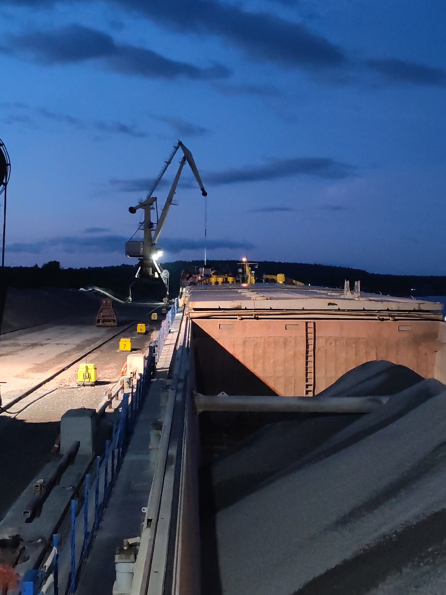 The image depicts a nighttime industrial scene at a port in Shalsky, Russia. A large cargo barge is docked, its deck piled high with dark, granular material, likely aggregate or coal. The barge's rusted hull is visible, contrasting with the lighter-colored deck. A loading crane, illuminated by spotlights, is positioned over the barge, with its boom extended. On the dock, several yellow markers numbered 5, 6, and 7 are visible, alongside other industrial equipment and barriers. The sky is a deep twilight blue with scattered clouds, and a dark treeline forms the background. The lighting suggests activity is ongoing despite the low light conditions.