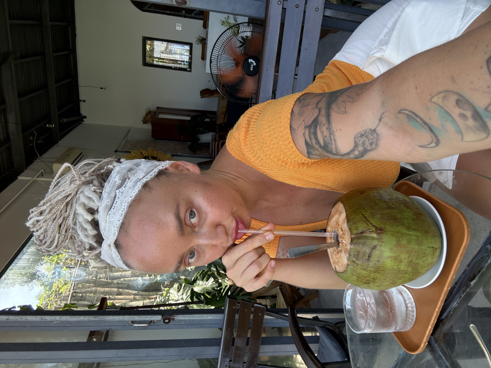 A woman with light-colored braided hair and a white lace headband is sitting at a table in an outdoor cafe in Bac Nha Trang Ward, Vietnam. She is wearing an orange top and white pants. In front of her is a whole green coconut with a straw and a spoon inserted, and a glass of iced water on a wooden tray. She is holding the straw to her mouth and looking at the camera. Behind her, a ceiling fan is visible, along with outdoor seating and lush greenery. The overall ambiance suggests a relaxed, tropical setting.
