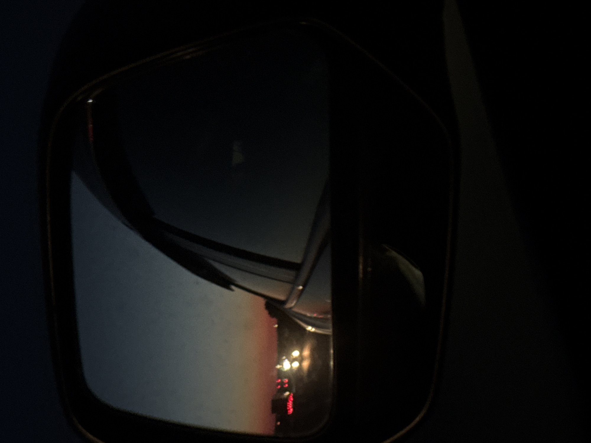 The image shows a car's side mirror at night. The mirror reflects a road with cars and traffic lights. The lighting suggests it's evening or nighttime. The reflection also captures a hint of the sky, which appears hazy or cloudy. The location is a city street with automobile traffic.