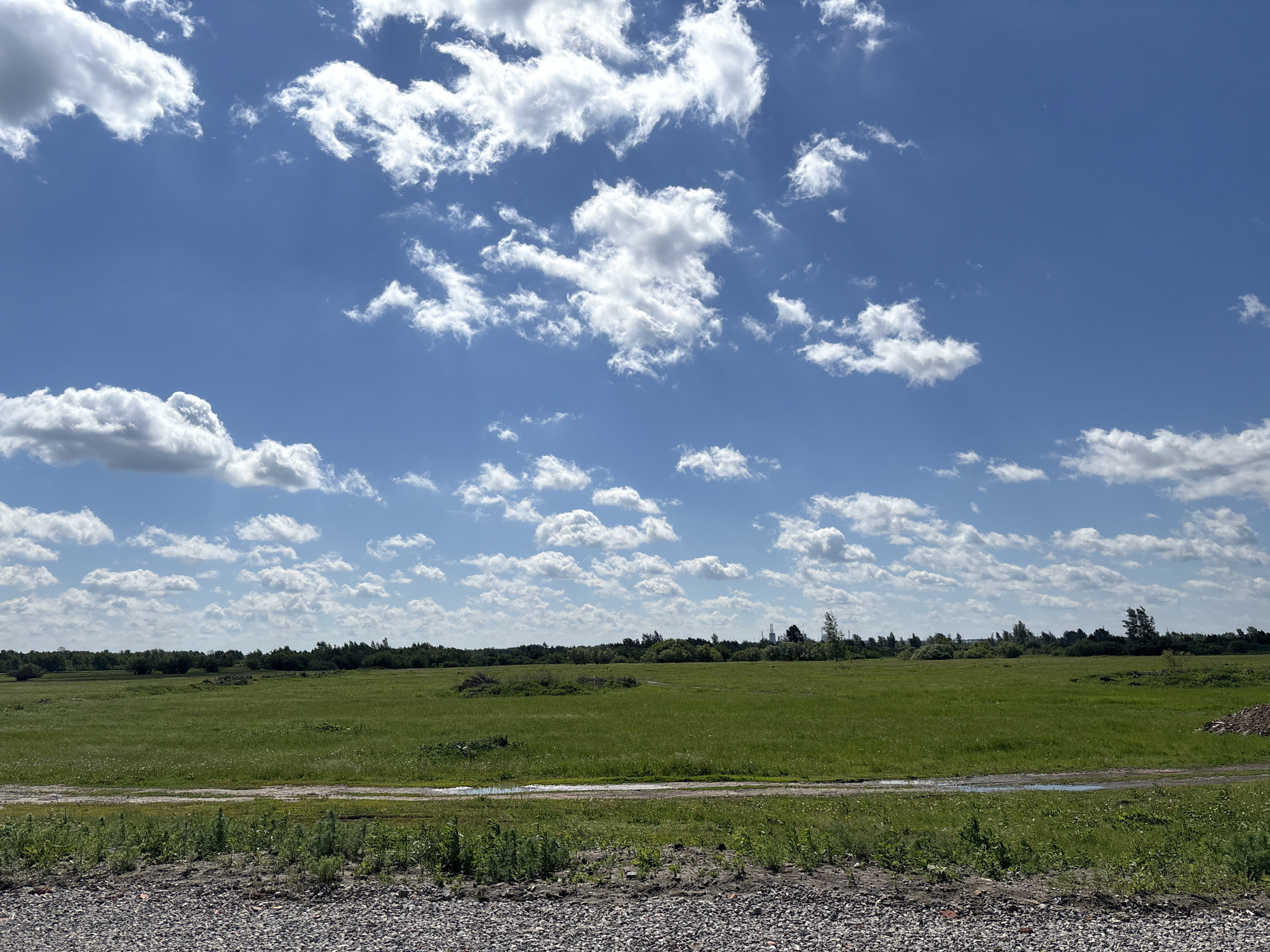 The image depicts a rural landscape in Ембаево, Russia. A wide, flat green field dominates the scene, stretching towards a distant line of trees. The sky is blue with scattered white cumulus clouds. In the foreground, there is a gravelly area or road. No people or specific subjects engaging in actions are visible. The environment is open and typical of a rural setting.