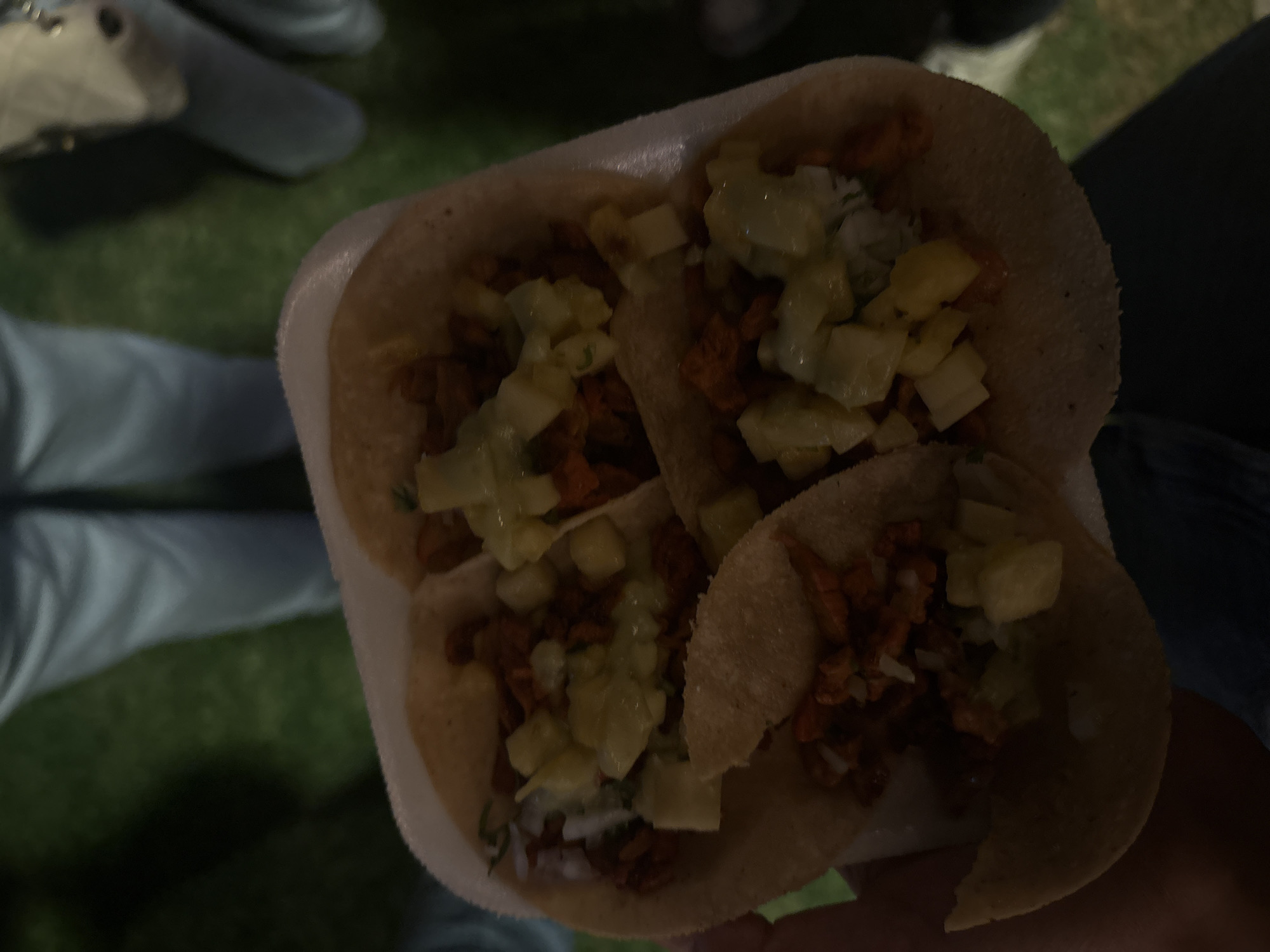 The image depicts a close-up view of three tacos al pastor served in a white styrofoam container. The tacos are filled with marinated pork, pineapple, and possibly onion. The background shows an out-of-focus view of a person's legs wearing light-colored pants and possibly a green grassy area. The image was taken in San Bernardino Tlaxcalancingo, Mexico.