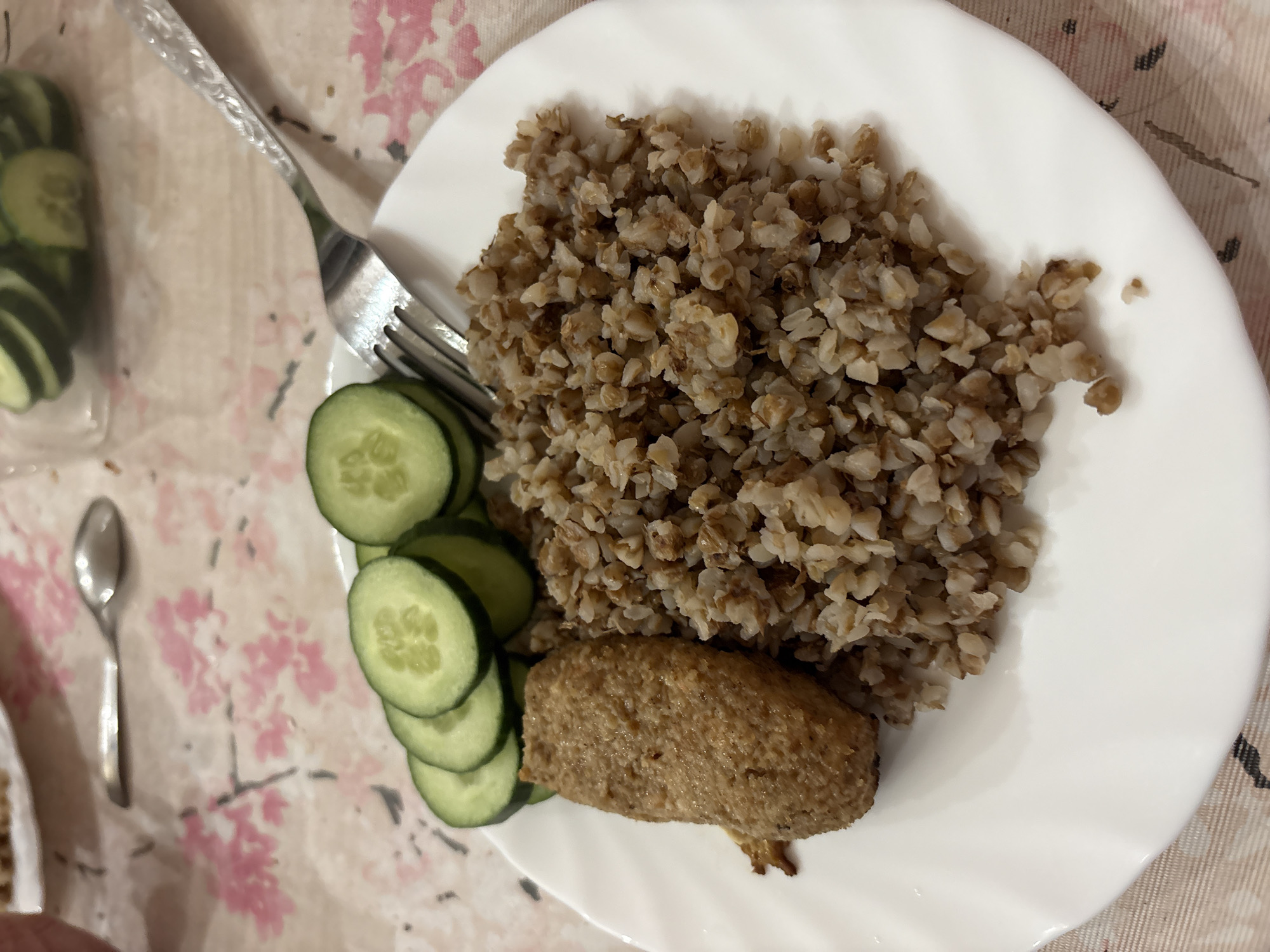 The image depicts a plate of food consisting of buckwheat, sliced cucumbers, and a meat patty, arranged on a white plate. A fork rests on the left side of the plate, near the cucumbers, with a spoon visible in the background. The plate is set on a tablecloth with a floral pattern. There is no indication of a specific location within the city of Berezniki, Russia, nor are there any people visible in the image.