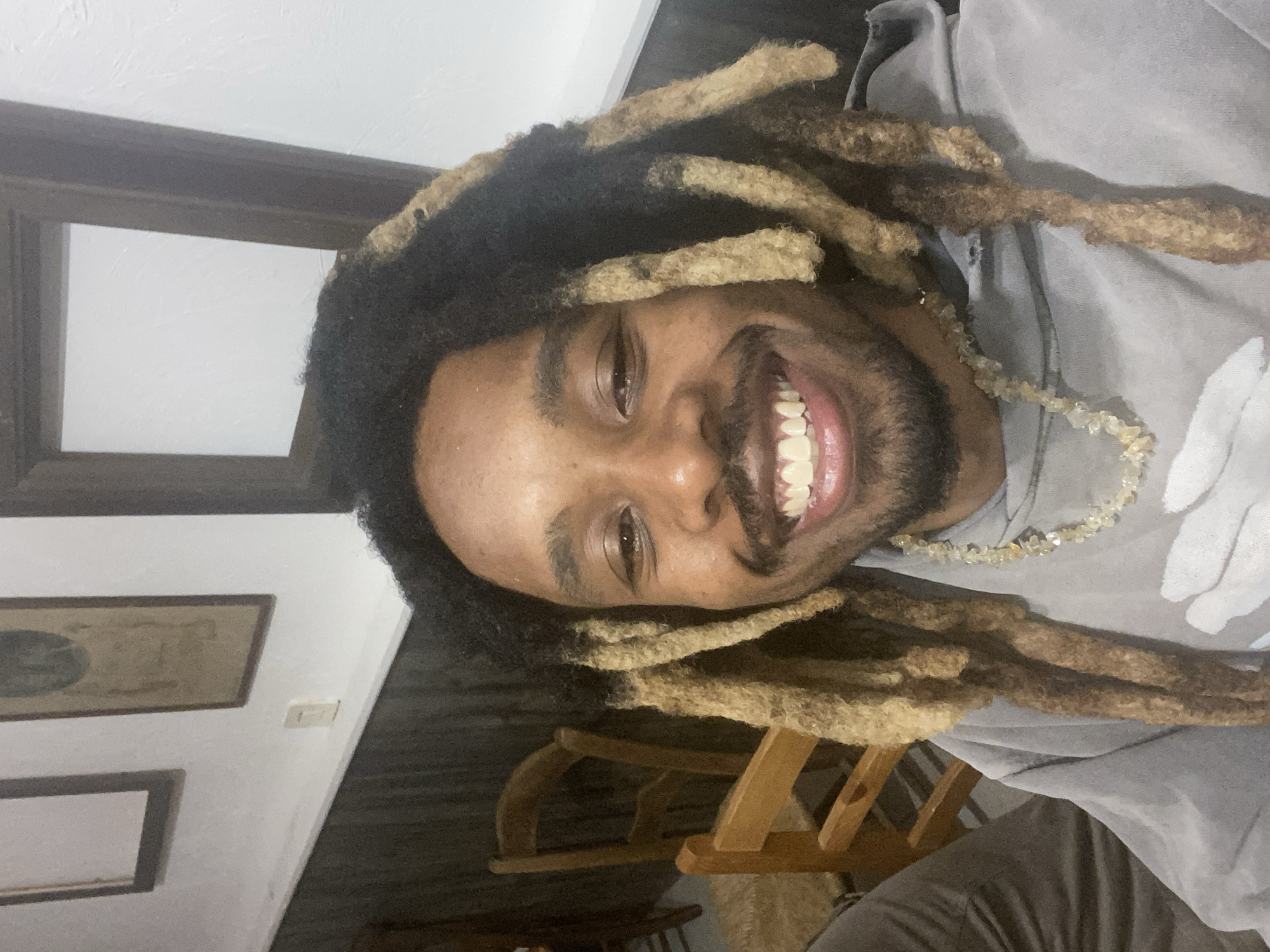 The image is a close-up, rotated selfie of a smiling young man in an indoor setting. He has dark skin, a prominent mustache, and a beard. His hair is styled in long dreadlocks, featuring a mix of dark and lighter, possibly blonde or light brown, colored sections. He wears a light grey hooded sweatshirt and a necklace crafted from light-colored, rough-cut beads. The man looks directly at the camera with a wide, cheerful smile, revealing his teeth and conveying a sense of happiness.
The background consists of white walls. Visible on the left side are dark wooden frames, likely for artwork or mirrors, including a smaller one below a larger one. A white light switch is also visible on the wall next to the frames. In the lower right, elements of wooden furniture, possibly parts of a chair, can be seen. The lighting suggests an indoor environment, and there is no discernible text in the image.