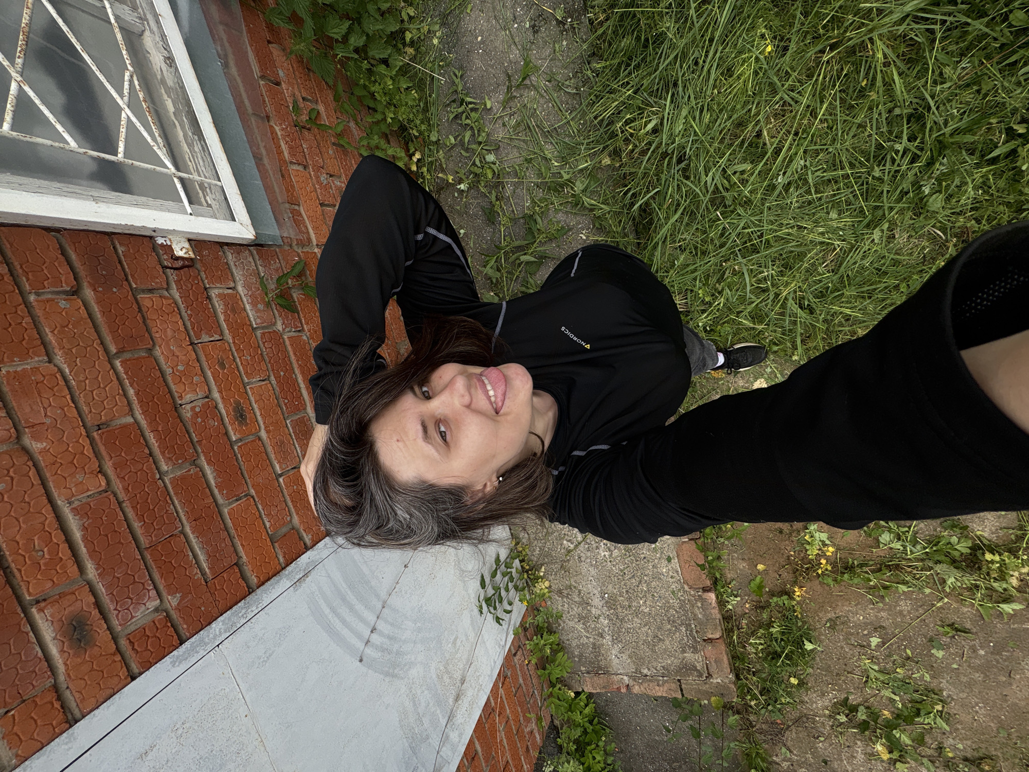 A young woman with brown hair and gray streaks is taking a selfie from a high angle in front of a brick wall. She is wearing a black long-sleeved shirt and black pants. She is sticking her tongue out and smiling. There is grass and concrete around her, and a metal door and a window with bars are visible in the background.