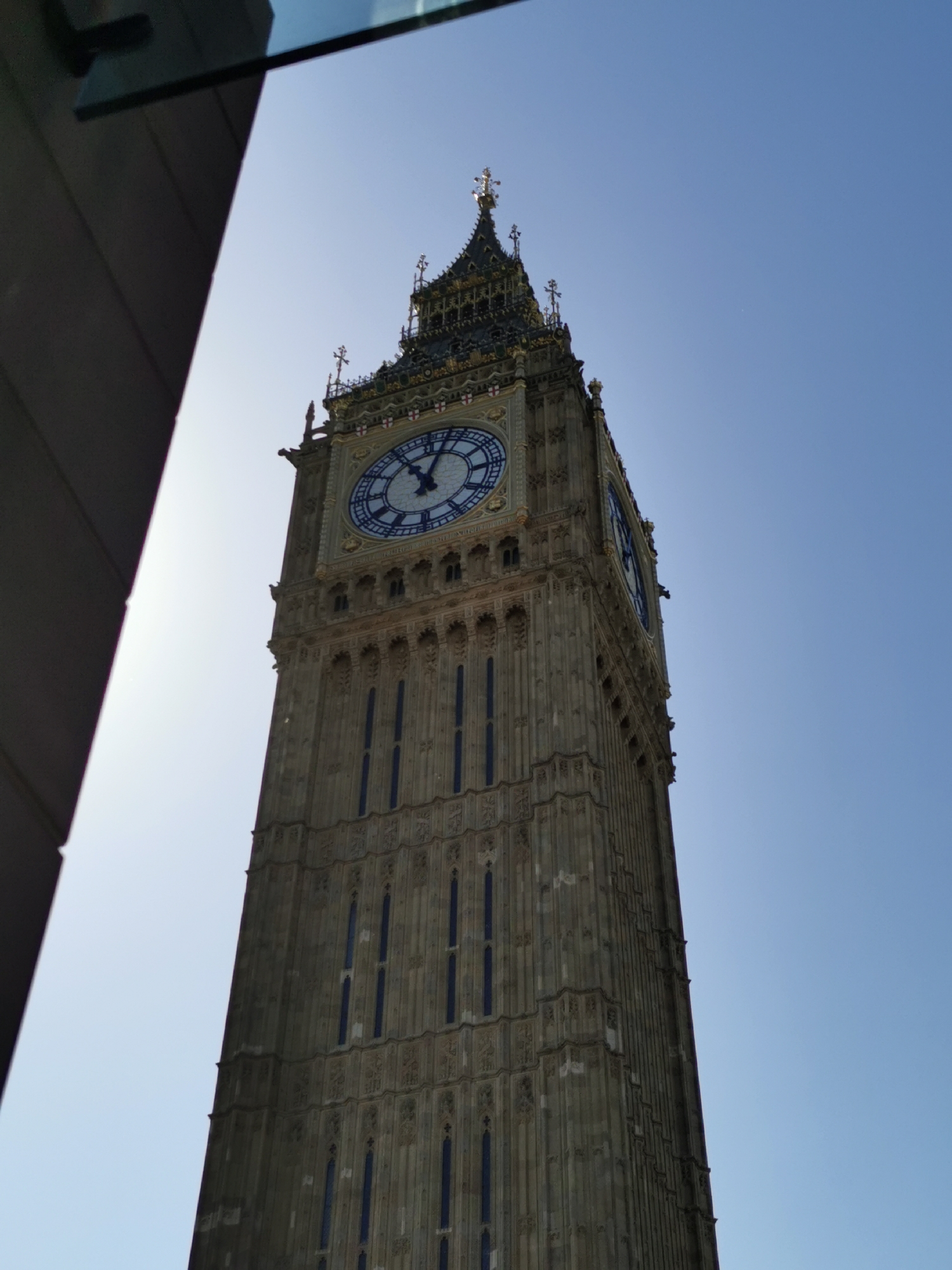The image depicts the Elizabeth Tower (commonly known as Big Ben) in London, United Kingdom. The tower is constructed of grey stone and features a large clock face with blue details. The sky is clear and blue. A building is visible on the left side of the image.