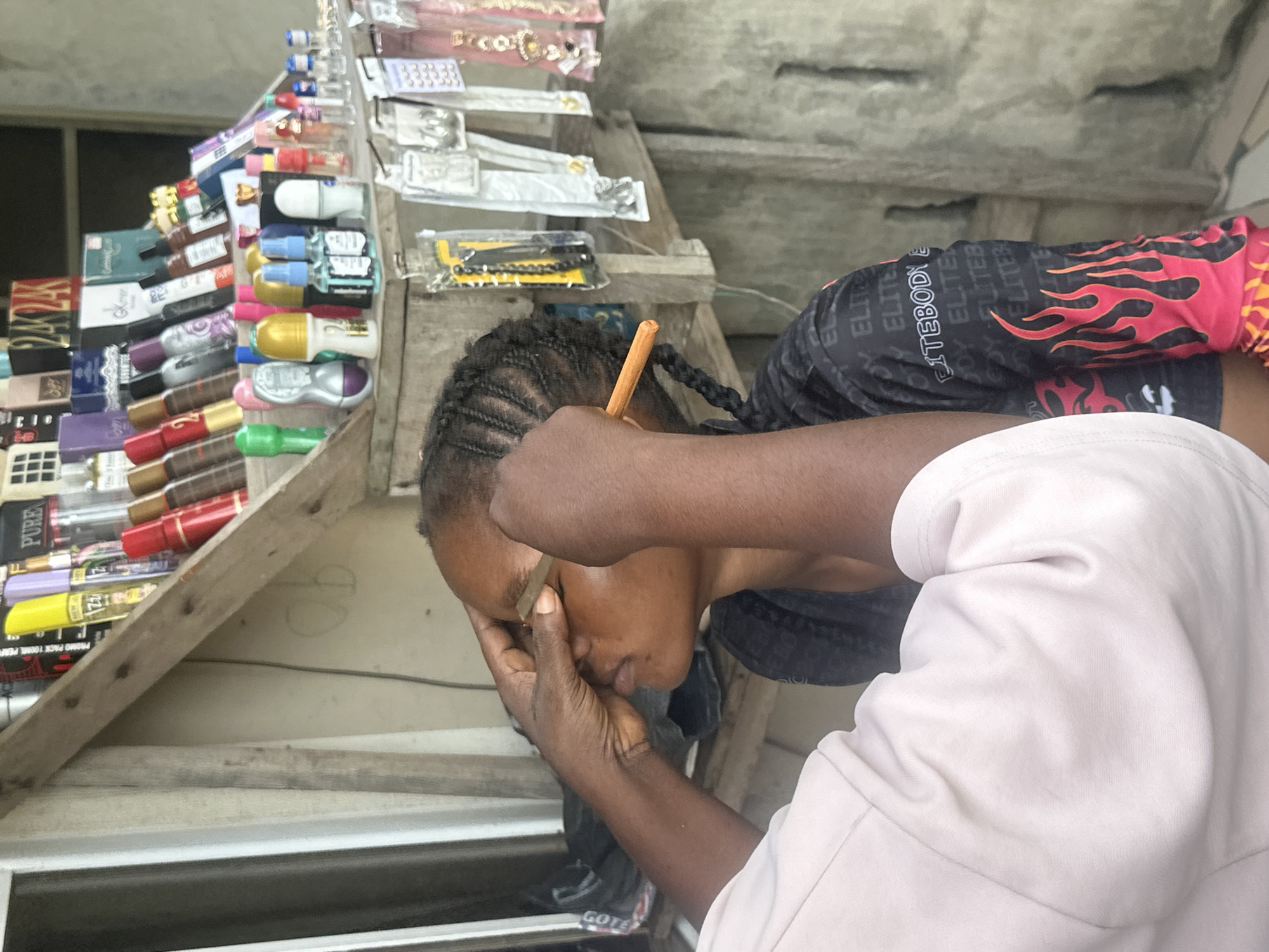 The image depicts a close-up of a woman's face as another person's hands are seen grooming her eyebrows with what appears to be a razor or eyebrow trimmer. The woman is of African descent with braided hair. She is wearing a black t-shirt with a flame design on the sleeve and the word "ELITEBODY" repeated across it. The person grooming her eyebrows is wearing a light pink garment.
The background suggests an outdoor market stall setting in Badagry, Nigeria. A wooden shelf is laden with various cosmetic products, including perfumes, deodorants, and hair accessories. The products are arranged in a somewhat haphazard manner, typical of a street vendor. The overall environment appears to be a local marketplace.