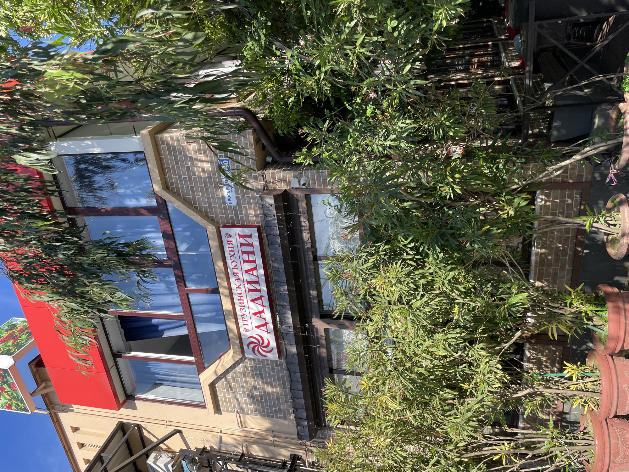 The image shows the exterior of a restaurant with Georgian cuisine called "Dadiani" located in Sochi, Russia. The restaurant has a red facade on the upper level and brick walls on the lower level. Large windows are visible, some of which are partially covered by blue curtains. In front of the restaurant, there are several potted plants with lush green foliage. A street sign indicating "просвещения 6" (Prosvenscheniya 6) is visible on the brick wall. The lighting suggests it is daytime, with sunlight casting shadows. There are no people visible in the image.