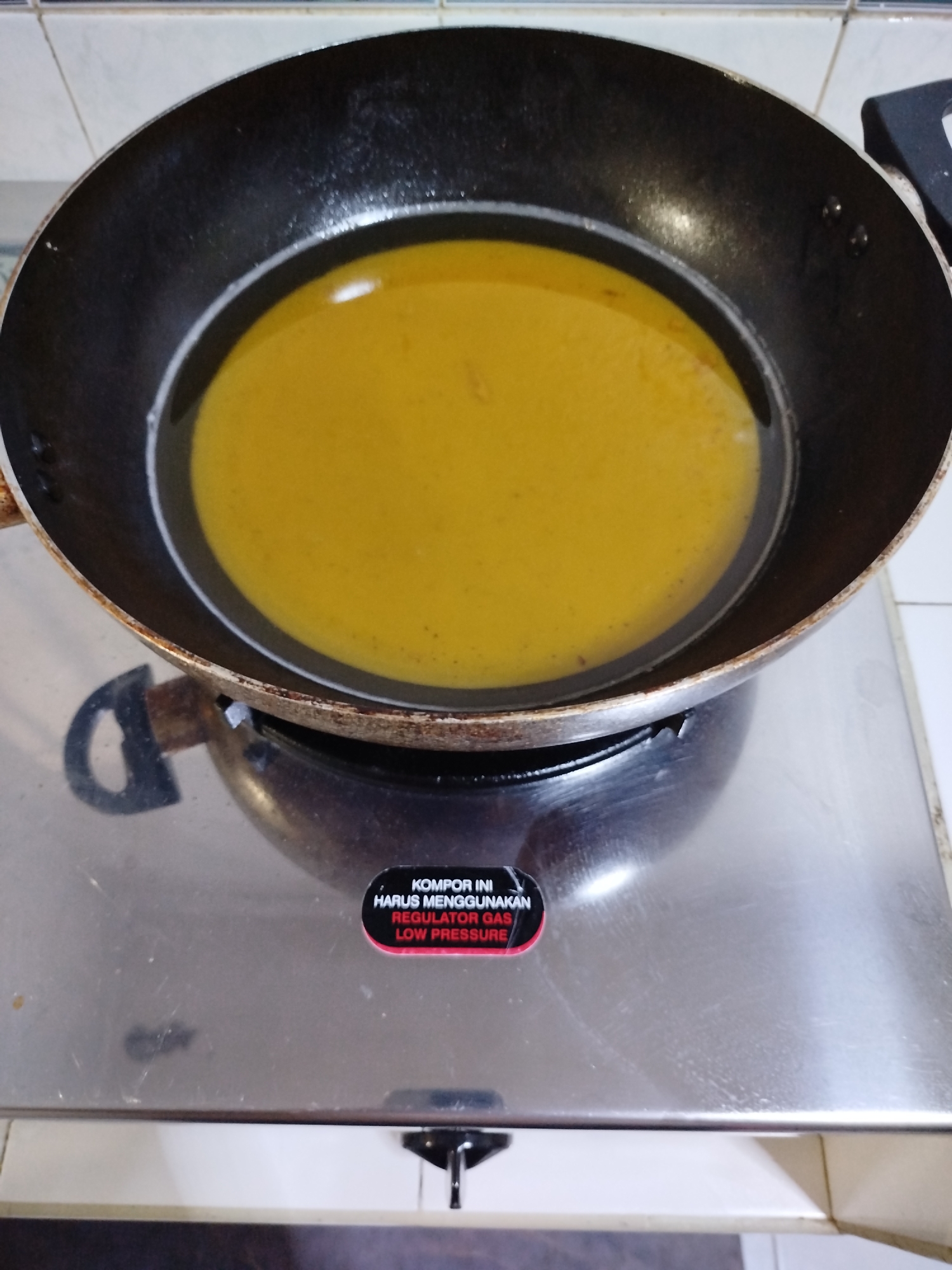 A black frying pan filled with hot yellow oil sits on a stainless steel stovetop. The stovetop has a black burner grate and a red and black sticker with Indonesian text. The background is a tiled wall. The scene is likely a kitchen in a home or restaurant.
