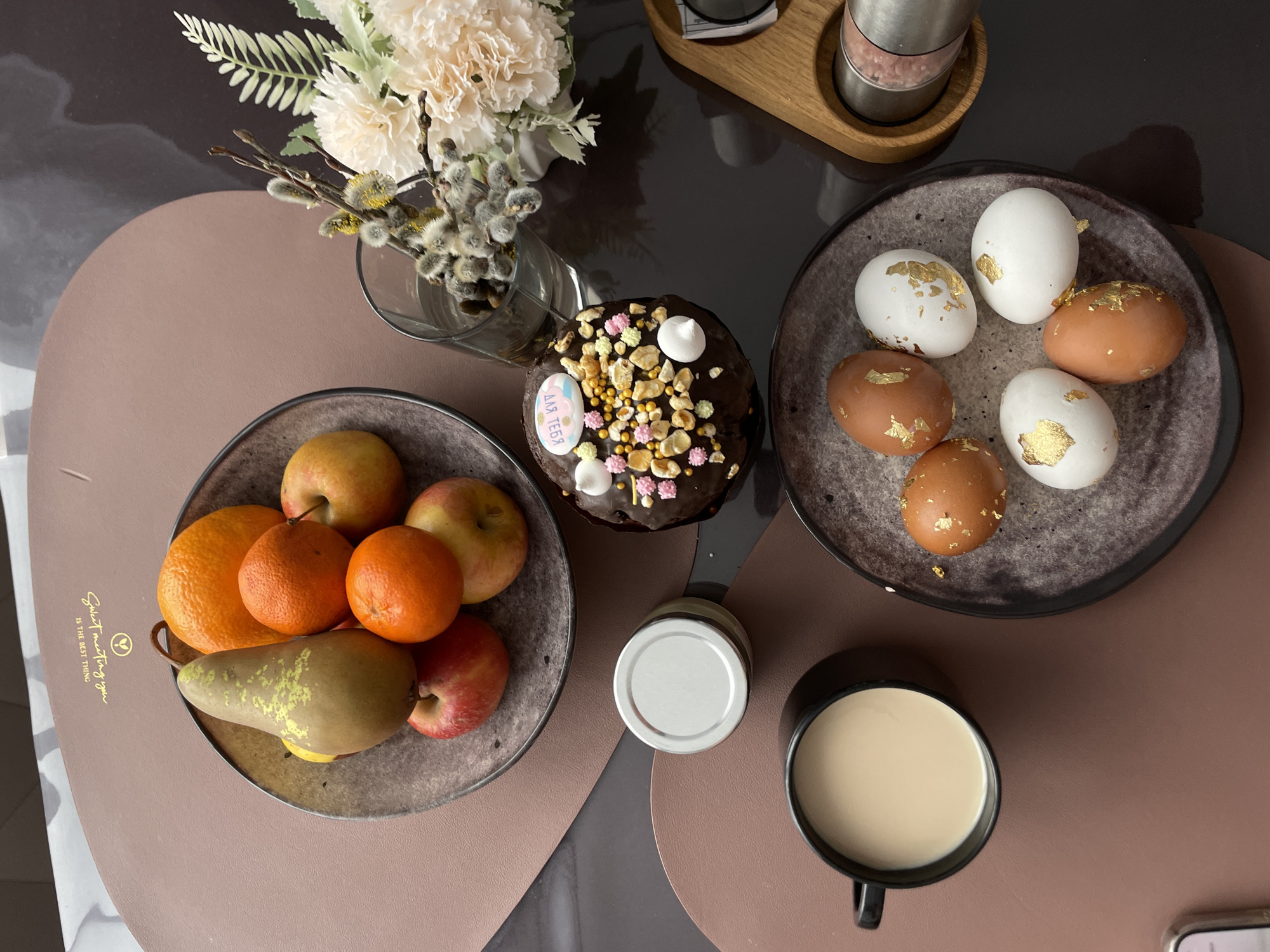 The image depicts an aerial view of a table setting. On the table, there are two dishes, one containing fruits (oranges, apples, and a pear) and the other with decorated Easter eggs. A decorated Easter cake is also present. There is a glass vase with flowers and pussy willows. A mug of coffee and a jar with a silver lid are placed nearby. The scene is likely indoors, given the lack of any specific identifiable details suggesting the exterior. The location is Подшивалово, Russia, though this information is not visually verifiable from the image itself.