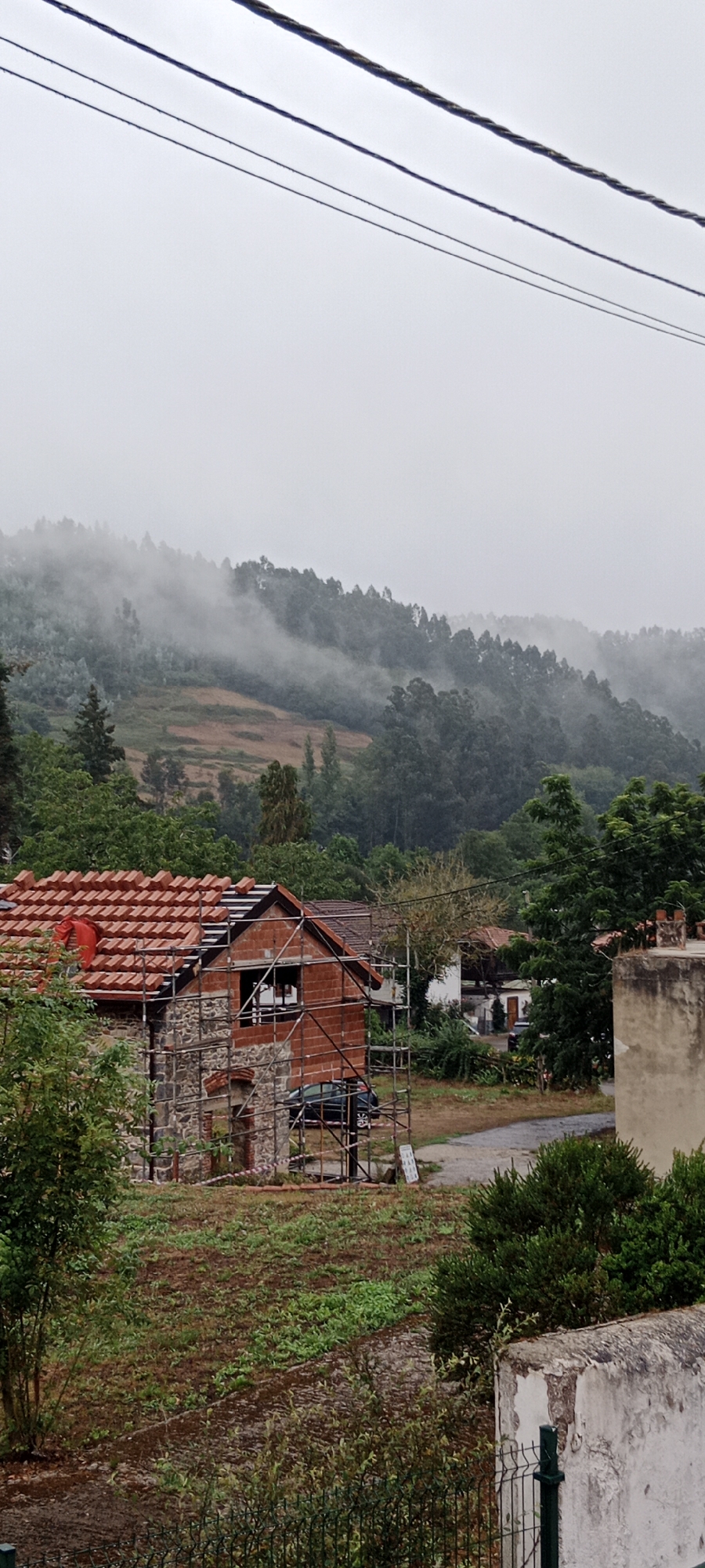 The image depicts a rural scene in Pravia, Spain, under an overcast sky. In the foreground, a house under construction is visible, with scaffolding surrounding it and red tarps covering part of the roof. The house features stone and brick construction. Adjacent to the house, there is a plot of land with uneven terrain and scattered vegetation. In the middle ground, other buildings and trees are present, and a dark-colored car is parked near the construction site. The background is dominated by a forested hillside, with mist or fog obscuring the upper parts of the trees, suggesting a damp or humid atmosphere. Overhead, electrical wires are strung across the frame.