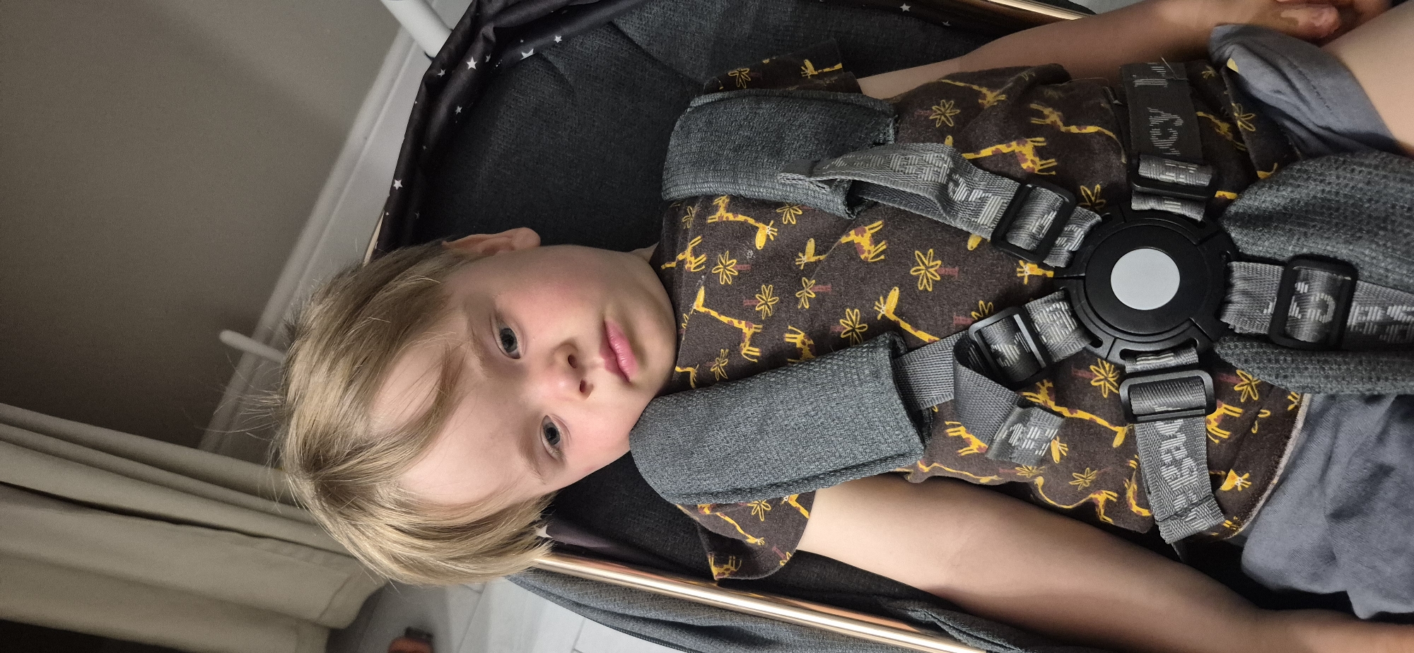 A blonde-haired child with blue eyes is lying on their back, looking towards the camera. They are wearing a brown t-shirt with a pattern of giraffes and flowers, and a grey harness with straps. The child is in what appears to be a stroller or some form of seating. The background is blurred, showing a light-colored wall and some draped fabric. The lighting suggests it is daytime, and the setting is indoors. The child's expression is neutral, with slightly rosy cheeks. There is no visible text in the image.