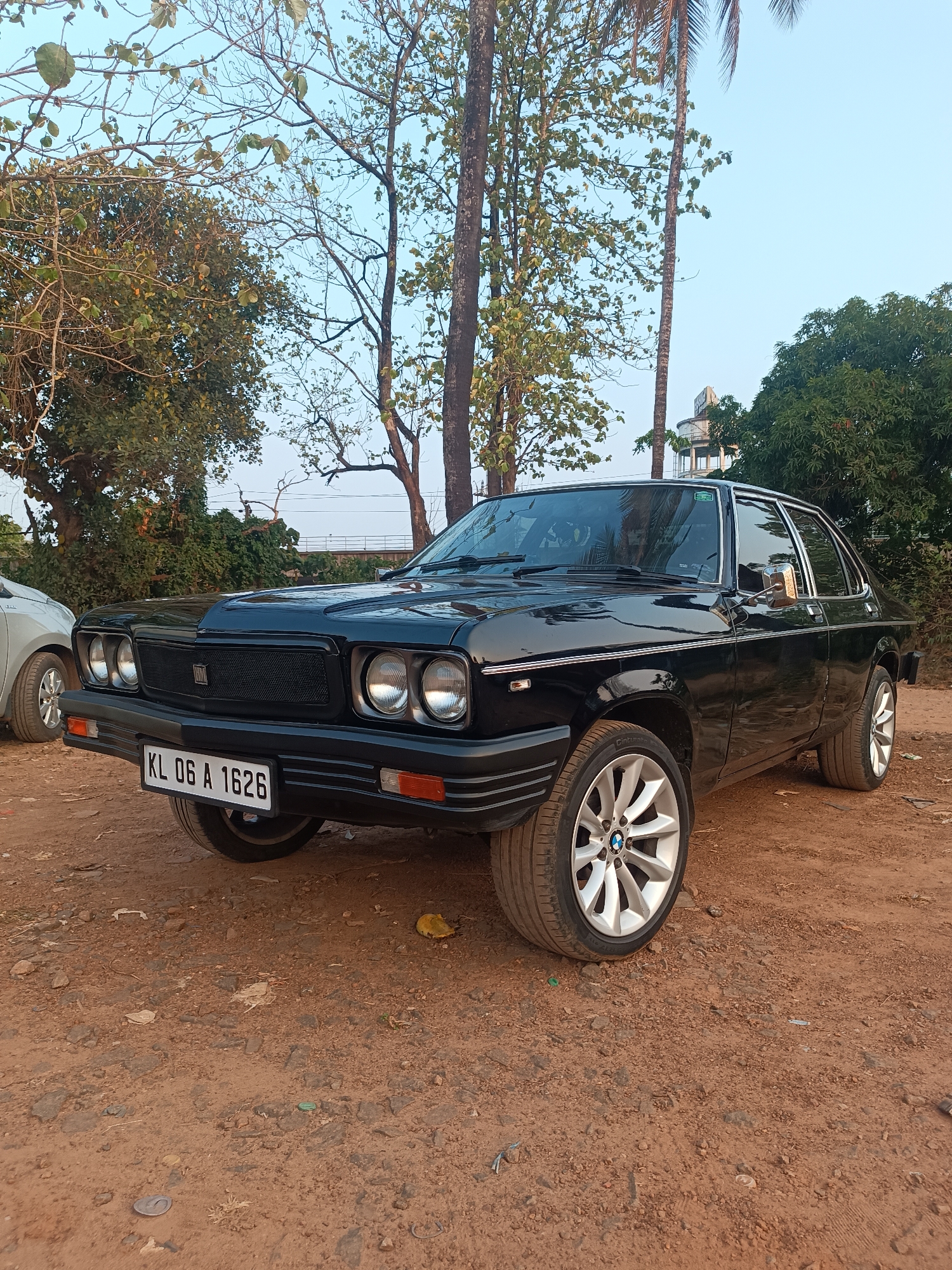 A black car with license plate "KL 06 A 1626" is parked on a dirt lot. The car has circular headlights and a mesh grill. A partially visible silver car is on the left. Trees surround the lot, and a water tower is visible in the background. The photo was taken in Kalamassery, India.