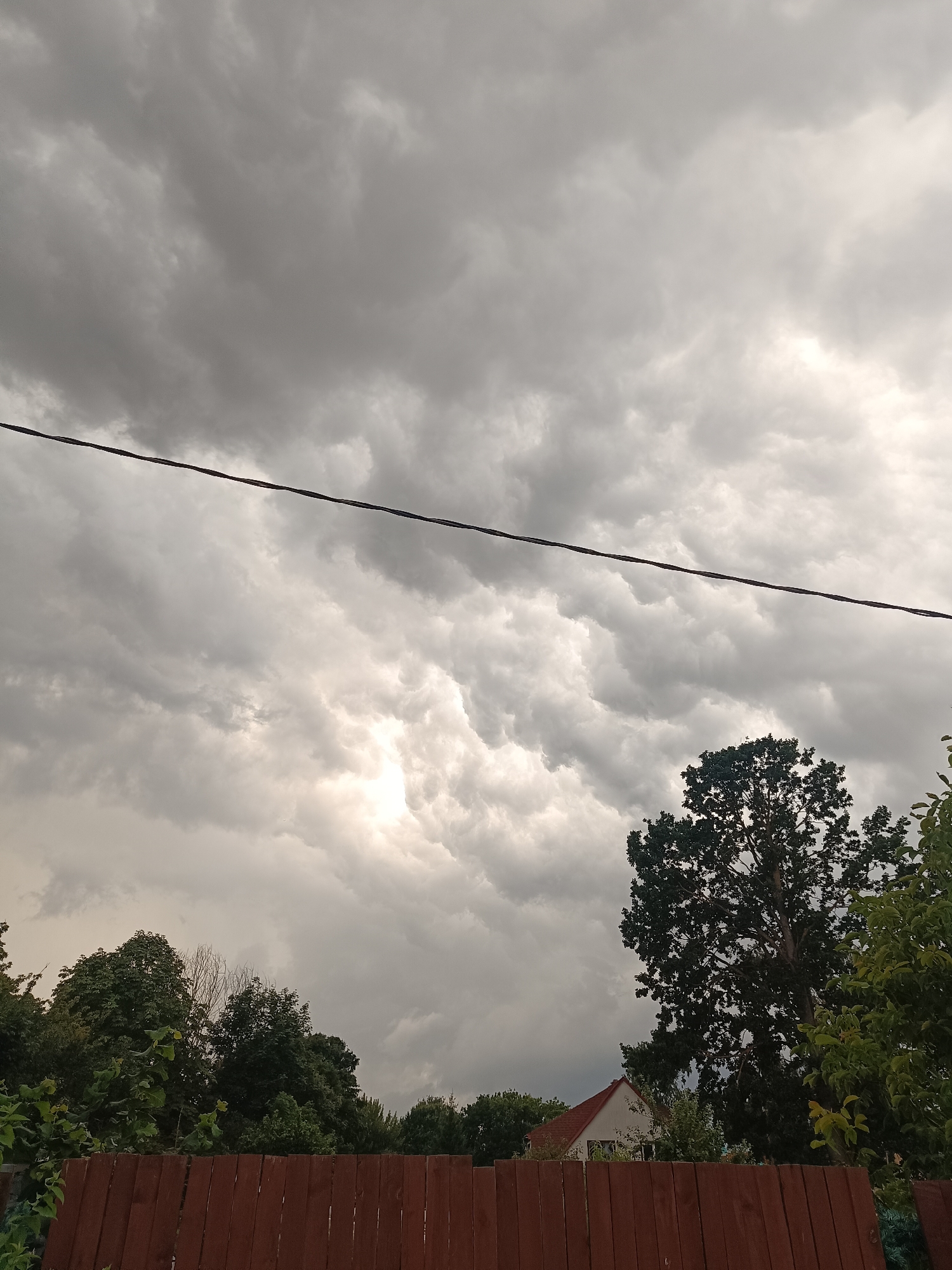 The image depicts an overcast sky filled with dramatic, heavy grey clouds. A dark, thin wire bisects the sky horizontally. Below the clouds, lush green trees are visible, with a larger, dark-leafed tree dominating the right side of the frame. A brown wooden fence runs across the bottom of the image, with a portion of a white house with a red-tiled roof peeking out from behind the trees on the right. The scene suggests impending rain or a recent storm. The location is identified as Riga, Latvia.