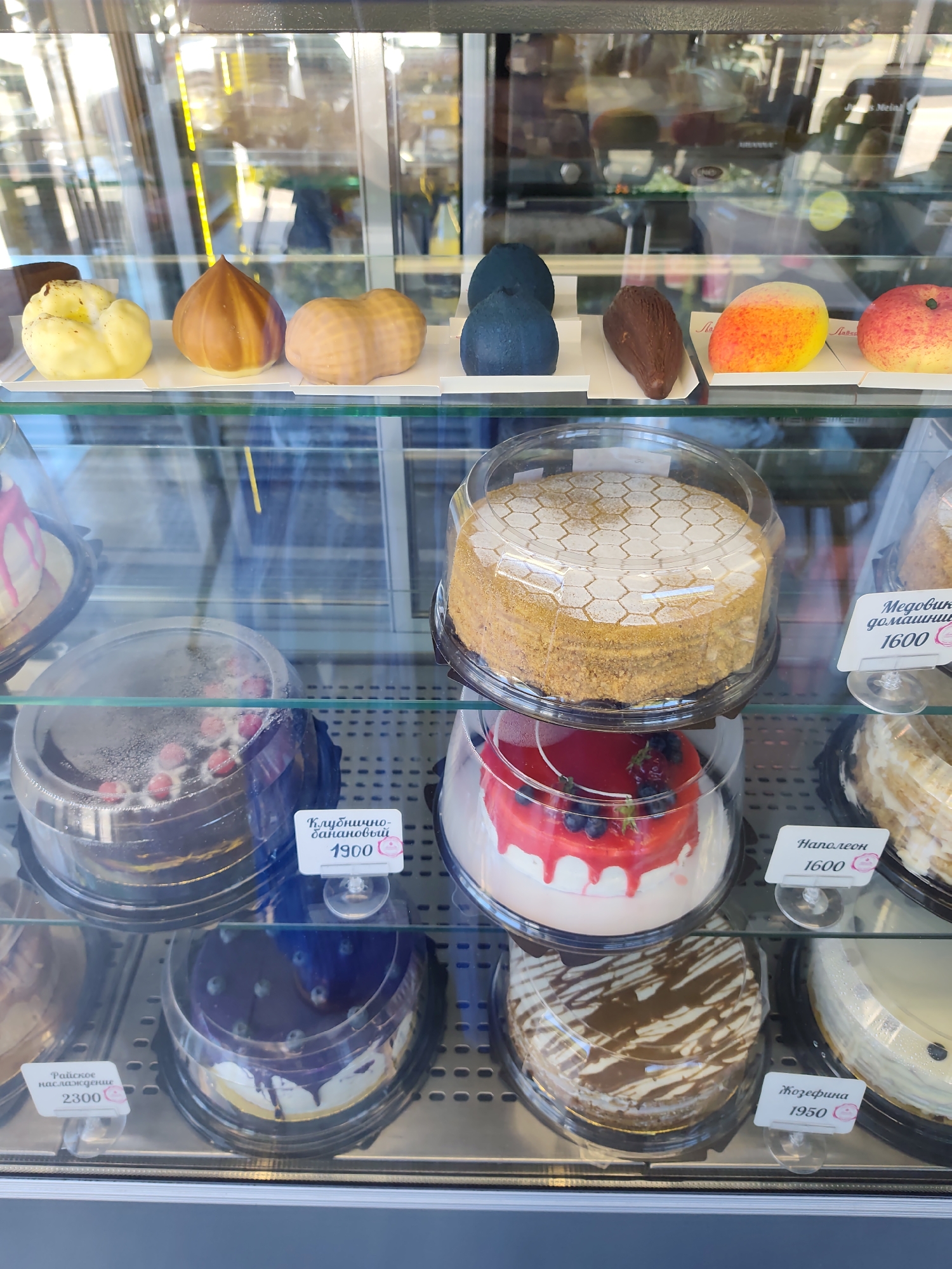 The image shows the interior of a brightly lit bakery display case in Sochi, Russia, filled with a variety of cakes and pastries. The display case is made of glass, reflecting the surroundings, including some yellow fluorescent lighting and what appear to be other shelves or display units. On the top shelf, several decorative pastries are arranged, some shaped like fruits and nuts, in colors ranging from yellow and orange to deep blue and brown.
Below this, various cakes are showcased. One prominent cake is covered with a honeycomb pattern and sits under a clear plastic dome. To its right, a cake labeled "Медовик домашний" (Medovik domashniy - Honey cake homemade) with a price of 1600 is visible. To the left of the honeycomb cake, another cake with red icing drizzled down its sides and topped with berries is displayed. Below that, a chocolate cake adorned with red berries and dusted with powdered sugar, labeled "Клубнично-банановый" (Klubnichno-bananovyy - Strawberry-banana) with a price of 1900, is visible. Further to the right, a layered cake labeled "Наполеон" (Napoleon) with a price of 1600 is present. On the bottom shelf, a cake with blue icing and berries is shown, along with a cake with a chocolate drizzle pattern, labeled "Жозефина" (Zhozefina - Josephine) with a price of 1950. Another cake, labeled "Райское наслаждение" (Rayskoe naslazhdenie - Heavenly delight) with a price of 2300, is partially visible on the far left. The overall scene depicts a well-stocked confectionery, presenting an array of sweet treats to customers. No people are directly visible in the image.