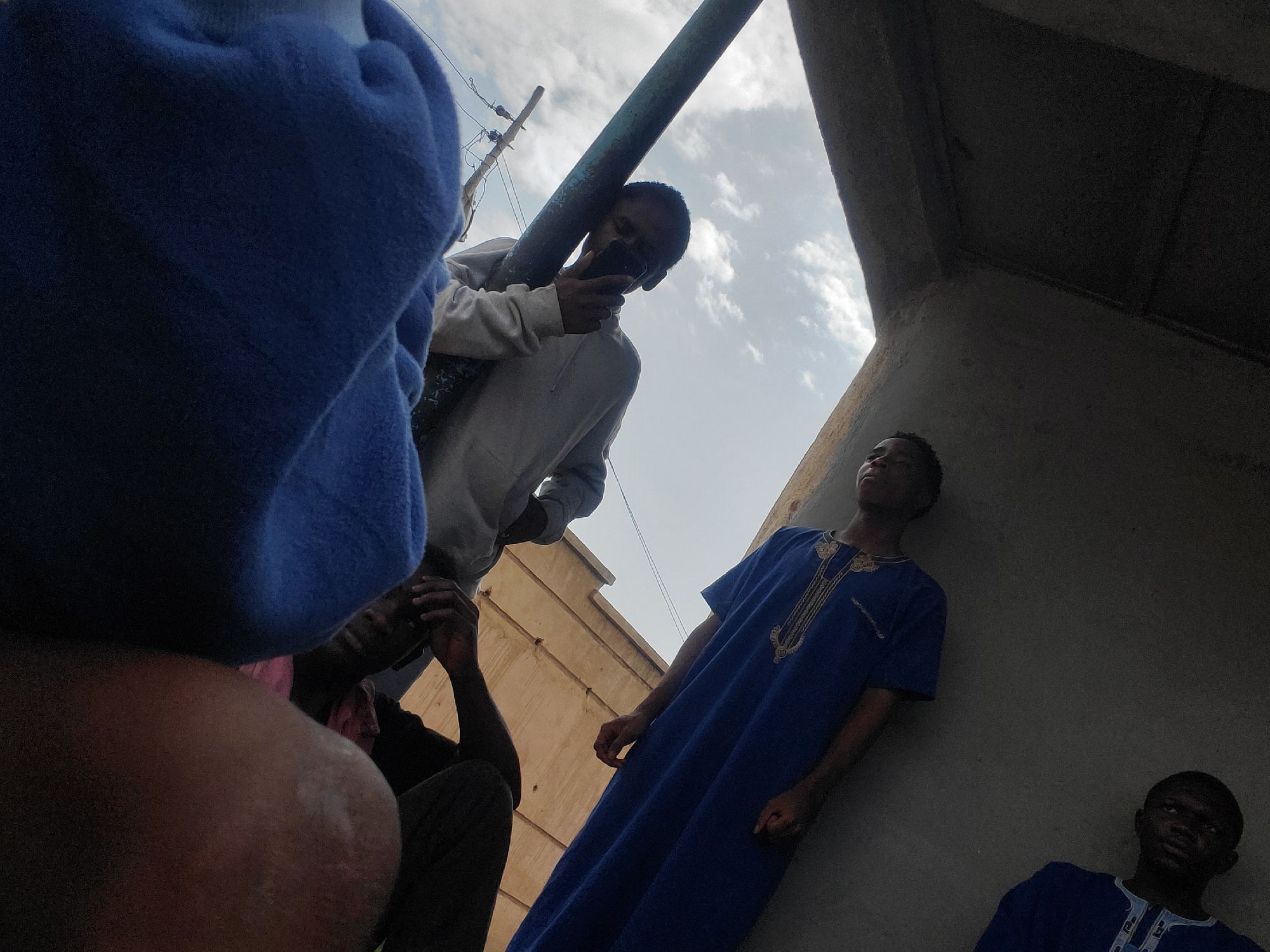 The image shows four people in an outdoor setting, possibly in the city of Gura Top, Nigeria. The perspective is from a low angle, looking upwards towards a concrete overhang and the sky. In the foreground, a person wearing a blue blanket is partially visible, with another person lying next to them, appearing to be looking at a mobile phone. A third person is leaning against a metal pole, also holding a phone. In the background, two individuals are standing, both wearing blue garments. The sky above is cloudy. The overall scene suggests a casual gathering in an urban environment.