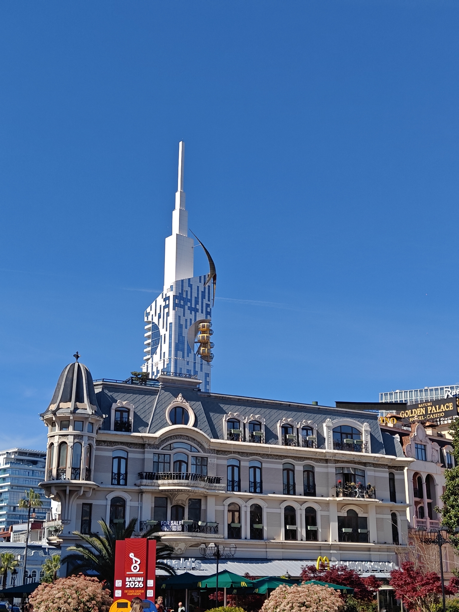 The image depicts a view in Batumi, Georgia, featuring a mix of architectural styles against a clear blue sky. Prominently displayed is a building with a classic European facade, characterized by ornate detailing, multiple floors, balconies, and a domed turret. Behind this building rises a modern, white skyscraper with blue patterns and a unique design, including what appear to be several partially exposed pod-like structures on its side.
At street level, the sign BATUMI 2026 EWF SENIOR EUROPEAN CHAMPIONSHIPS is visible along with the McDonald's restaurants. Part of the BATUMI GOLDEN PALACE HOTEL-CASINO sign can also be seen above the classic european building. The presence of trees, shrubs, and a sign indicates an urban street scene. Some pedestrians can be seen walking in the front of the picture.