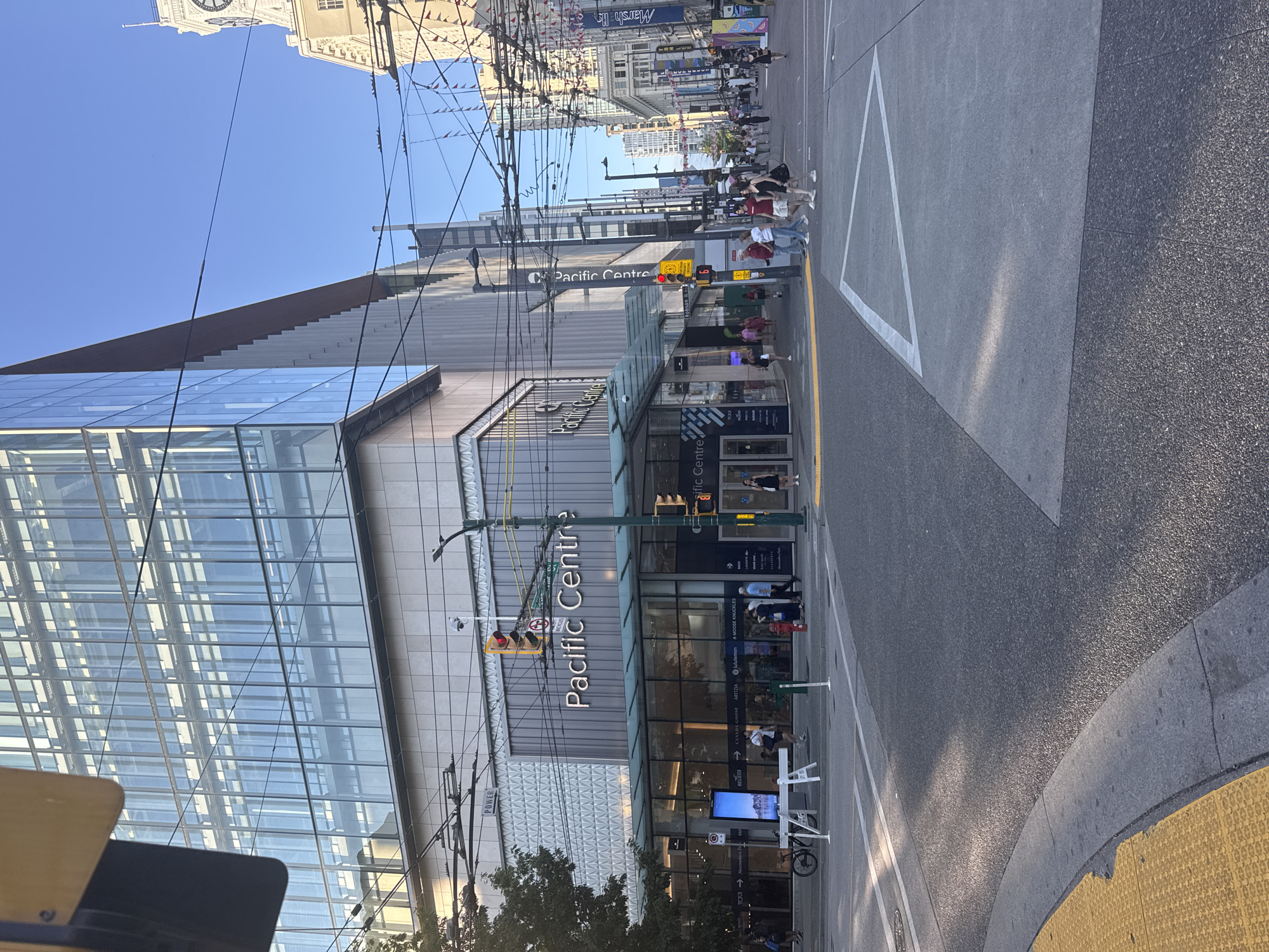 This image captures a sunny afternoon street scene at Pacific Centre in downtown Vancouver, Canada. A modern, multi-story glass building dominates the left side of the frame, its facade reflecting the clear blue sky. Below the glass structure, the entrance to Pacific Centre is visible, with people entering and exiting the shopping complex. A large sign prominently displays "Pacific Centre."
The street in the foreground is paved with asphalt and features a yellow tactile warning surface at the curb. Several people are walking on the sidewalks and crossing the street. Traffic lights are visible, and overhead wires suggest an active transit system. In the background, other buildings and a clock tower can be seen, indicating a bustling urban environment. The overall impression is one of a vibrant and active city center on a pleasant day.
