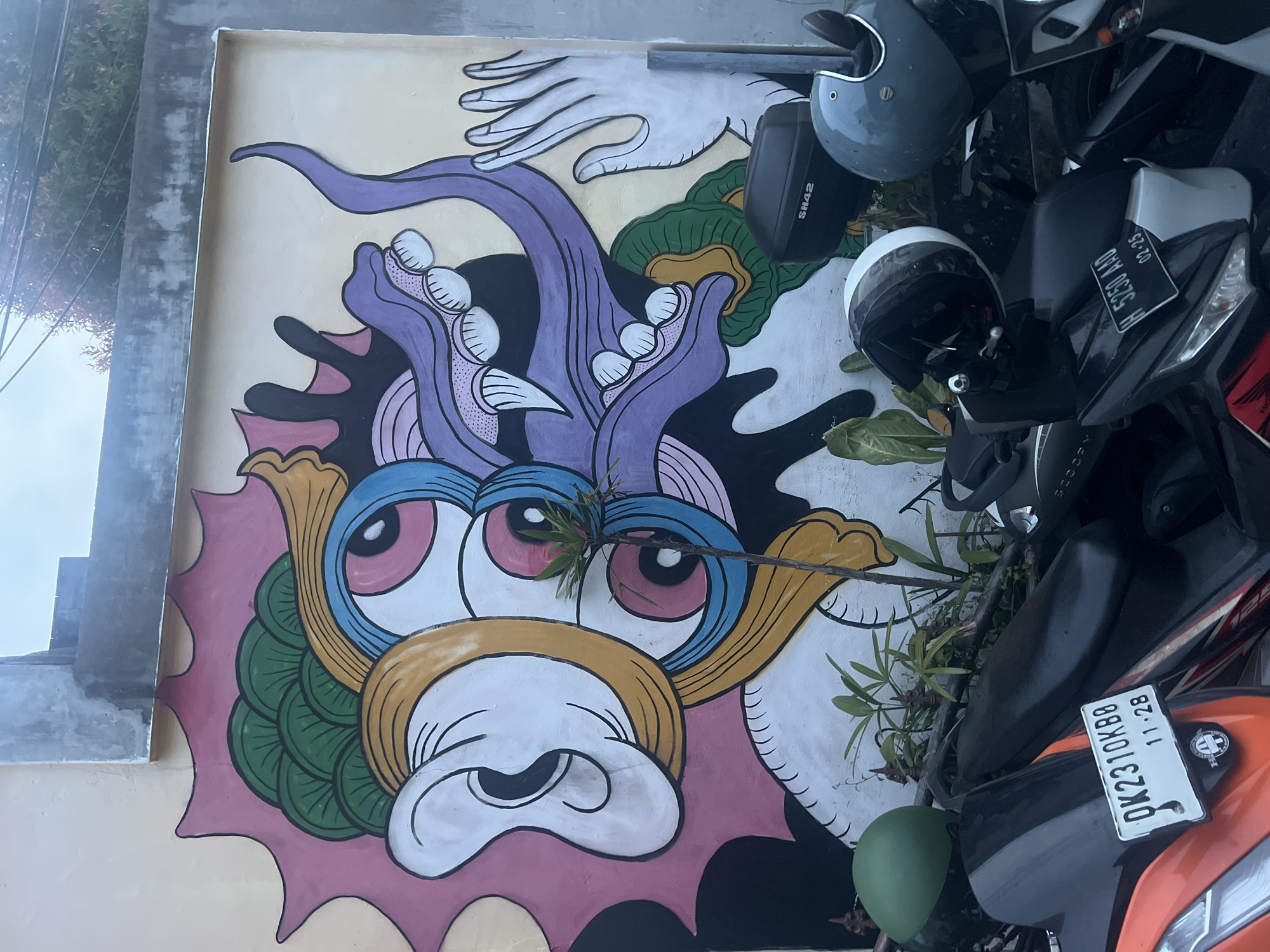 The image depicts a colorful mural on a wall in Tibubeneng, Indonesia, partially obscured by parked motorbikes and helmets. The mural features a stylized, cartoonish face with large pink eyes, a bulbous white nose, and a purple and black headdress. Above the head, there is a disembodied hand. Two motorbikes are parked in front of the mural, partially obscuring the lower right portion of the artwork. One bike is orange and black with the license plate "DK2310KBO". The other motorbike is grey and black with the license plate "H5250AAD". Two motorcycle helmets, one grey and one white and black, sit on top of the motorbikes and near the mural. There are also some plants growing near the base of the wall in front of the mural.