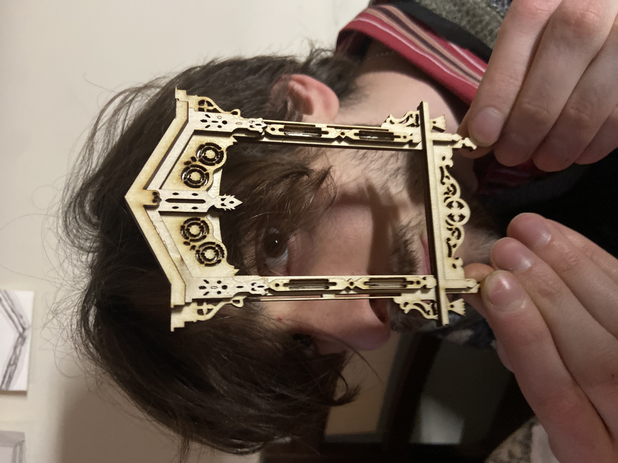 A person with brown hair and a beard is holding a wooden laser-cut frame in front of their face. The frame is intricate, with geometric and floral designs. The person's eye is visible through the frame. The background is blurry, but a wall and part of a painting can be seen. The scene is indoors. There is no indication that the photo was taken in Moscow, Russia.