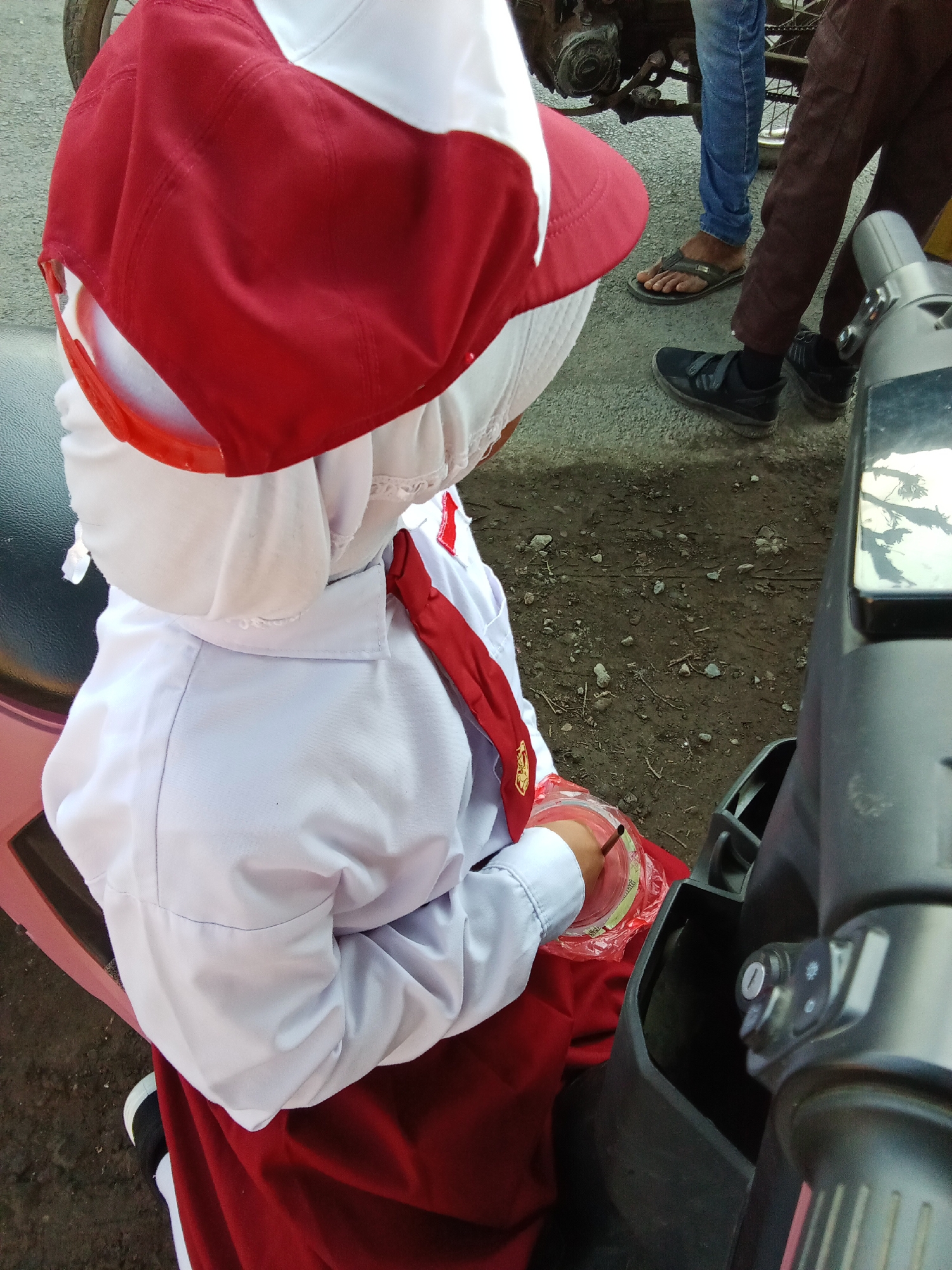 In Bungatan, Indonesia, a young student, viewed from behind, is seated on a pink scooter. They are wearing a white shirt, a red tie, a red and white cap, and a red skirt. The student's left hand is holding a clear plastic cup with a dark straw, resting on their lap. The background shows a dirt and gravel ground with some debris. In the upper background, two adults are partially visible: one in blue jeans and flip-flops, and another in brown trousers and black shoes. The scooter's handlebars and front panel are in the foreground on the right side of the image.