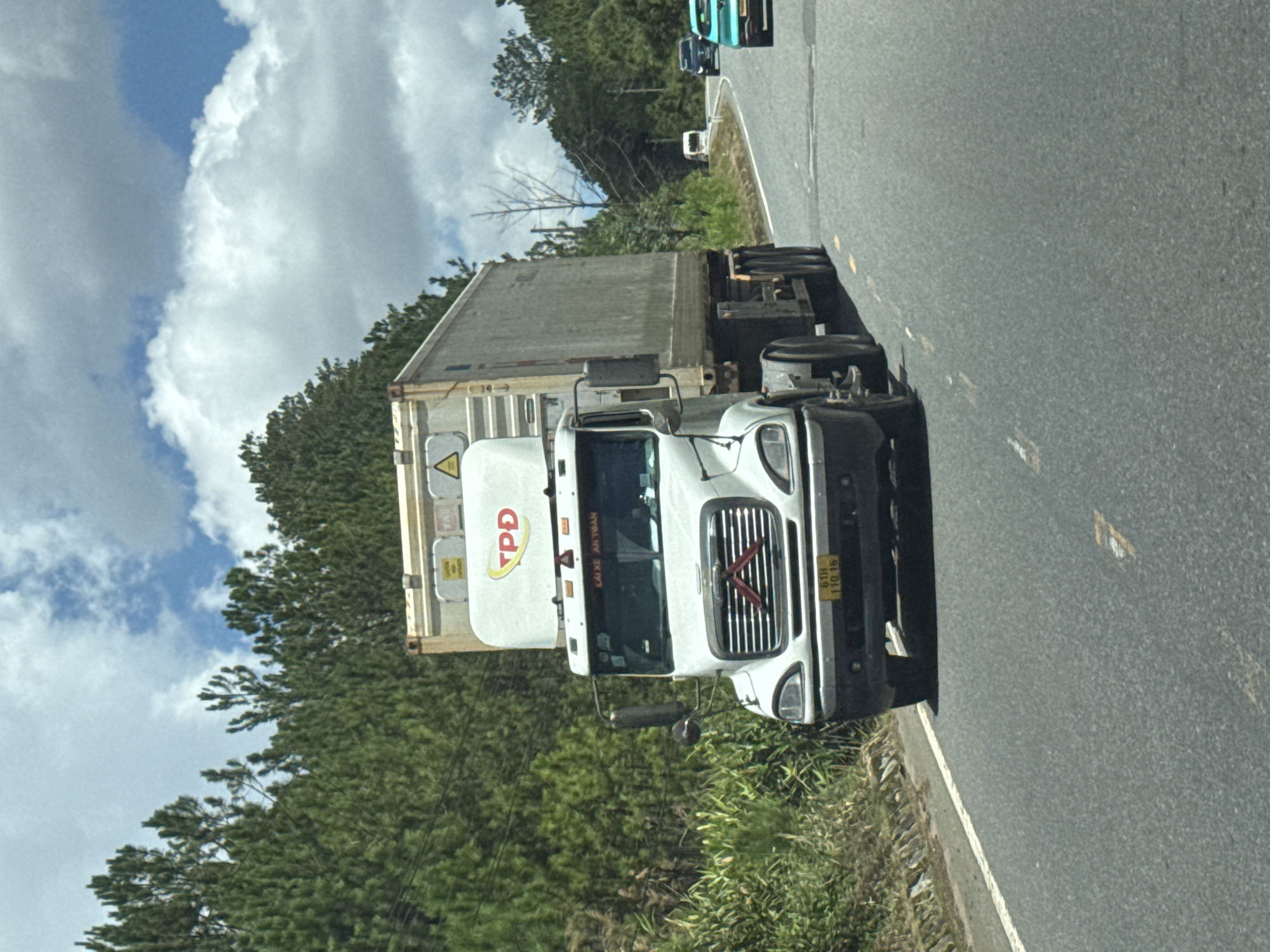 A white semi-truck with a grey shipping container is traveling on a paved road in Thôn 6, Vietnam. The truck is in the foreground and takes up a significant portion of the frame. It has a "TPD" logo on the side of the cab. The road appears to be a two-lane highway with a white dashed line marking the center and a solid white line on the right shoulder. Green trees and vegetation line the left side of the road. In the background, there are more trees on rolling hills, and a cloudy blue sky. Other vehicles, including a black car and a teal-colored car, are visible further down the road. The environment appears to be rural or semi-rural.