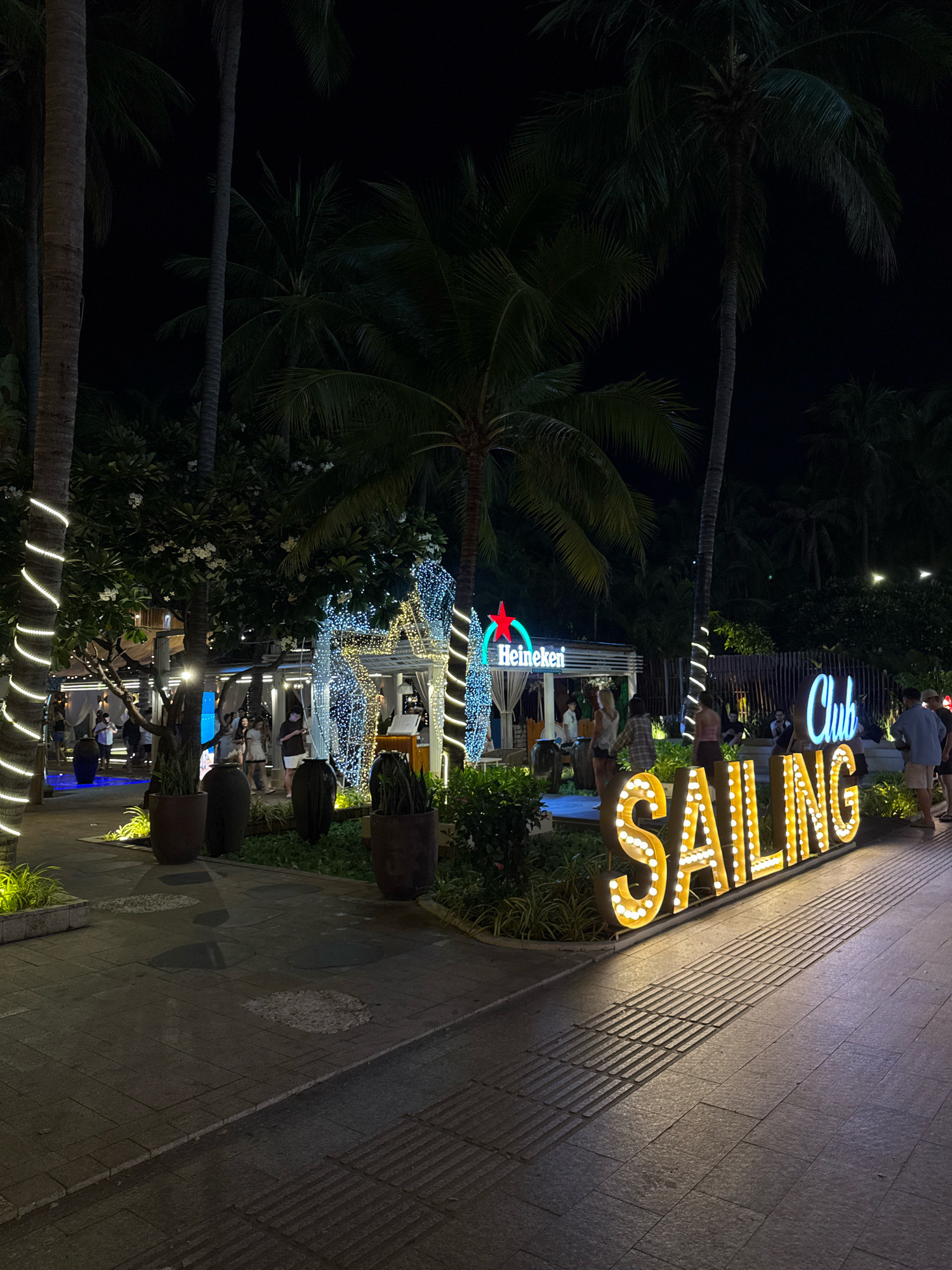 This image depicts a nighttime scene in Nha Trang, Vietnam, likely near a coastal promenade or entertainment district. Dominating the foreground is a large, illuminated sign spelling out "SAILING" in prominent, warm-toned light bulbs. To the right of the "SAILING" sign, a smaller, blue neon sign reads "Club."
Behind the signs, a building with open-air seating and white tents is visible, adorned with festive blue and yellow lights that outline structures and figures. A prominent "Heineken" logo with a red star is also visible above the entrance of this establishment.
Several people are present in the scene, mostly clustered around the building or walking along the illuminated pathway. They are casually dressed, suggesting a relaxed evening atmosphere. Palm trees with some trunks wrapped in white string lights frame the scene, adding to the tropical ambiance. The ground is paved with dark stone tiles, and decorative potted plants and planters are scattered throughout. The overall impression is one of a lively and illuminated nighttime entertainment venue.