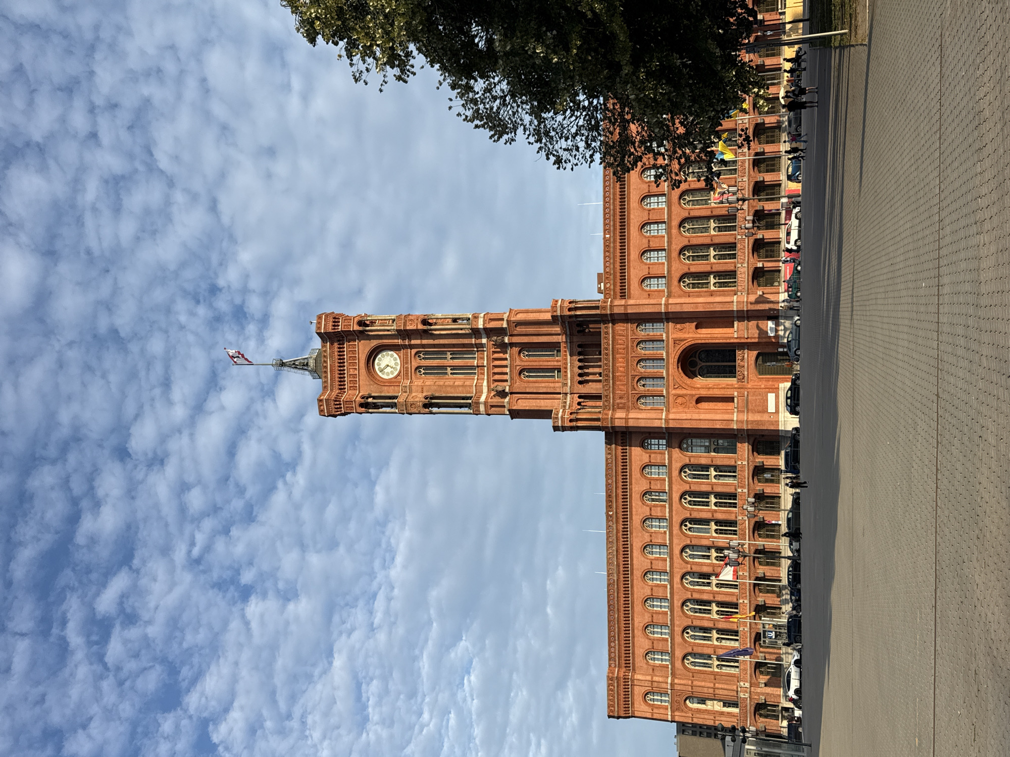The image shows the Rotes Rathaus (Red City Hall) in Berlin, Germany, under a partly cloudy sky. The building is constructed of red brick and features a prominent clock tower topped with a flagpole flying a German flag. The main facade of the building is symmetrical with numerous arched windows. In front of the building, a large paved plaza is visible, with several cars parked along the street and a few people walking. A large tree with green foliage is present on the right side of the frame. The lighting suggests it is daytime, likely late afternoon, given the long shadows cast on the plaza.