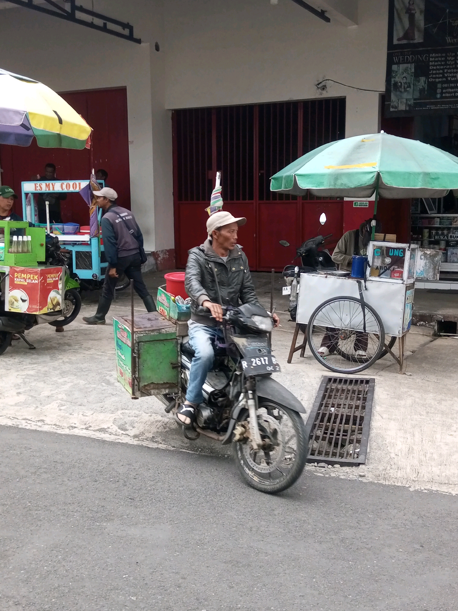 A man is riding a motorcycle with a sidecar towards the viewer in Krasak, Indonesia. He is wearing a grey jacket, blue jeans, and a white hat. The motorcycle is dark gray and has a license plate that reads "R 2617 B". The sidecar is green and has a white and green sign on it. The man is looking to his right. There is a food cart with a green umbrella in the background to the right, and another food cart with a yellow and purple umbrella to the left. There are also several people standing around the food carts. The ground is concrete and there is a metal grate in the foreground. The buildings in the background are red and white.