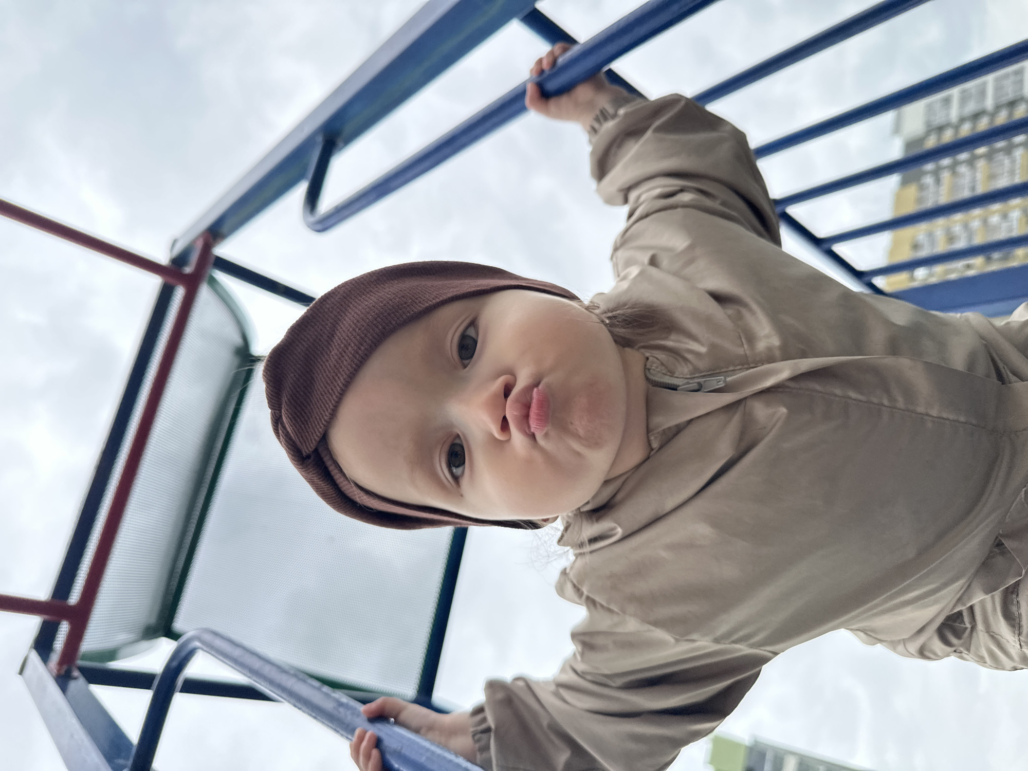 A low-angle, close-up shot shows a young child wearing a brown beanie and a beige jacket. The child is holding onto blue metal bars of playground equipment and puckering their lips. In the background, a cloudy sky and residential buildings are visible. The image was captured in Yekaterinburg, Russia.