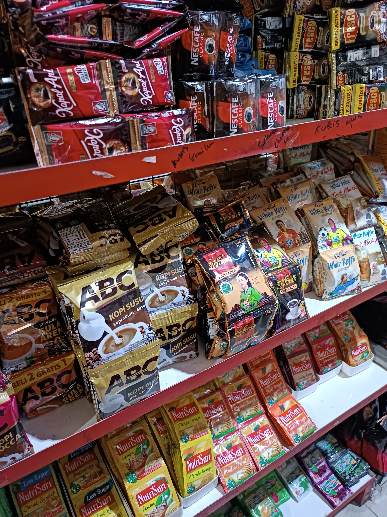 The image shows a shelf in a small convenience store in Jetis, Indonesia, filled with various coffee and beverage packets. The items are densely packed, with multiple brands and varieties displayed. Prominent brands include Nescafe Classic, Kapal Api, ABC Kopi Susu, and NutriSari. The shelves are made of red metal with wire racks. Some packets are stacked vertically, while others are laid flat. There are handwritten labels in Indonesian on the shelves, such as "kopi kurus" (skinny coffee). The lighting is a mix of ambient store lighting and possibly overhead lights, creating some glare on the packaging. The overall impression is of a well-stocked, local retail environment.