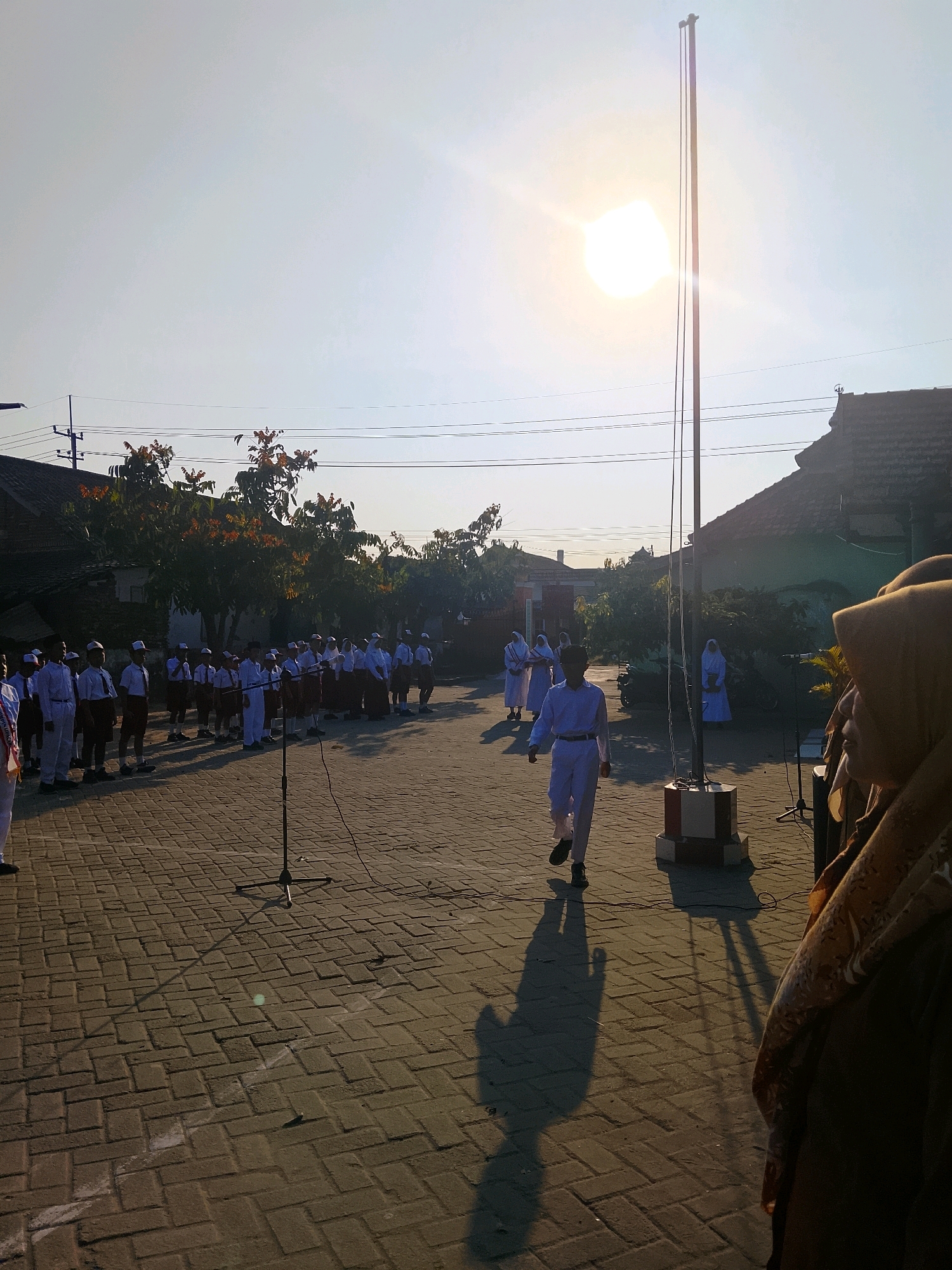 This is a medium-shot, eye-level, vertical, outdoor photograph of a flag-raising ceremony at sunrise in Kepuh, Indonesia. The sun, a bright disc, is positioned high in the sky, casting a strong glare and long shadows across the paved courtyard. A tall flagpole stands in the center of the frame, with two dark ropes running parallel down its length.
A group of students, dressed in white uniforms and caps, are lined up on the left side of the frame, facing the flagpole. They appear to be participating in a formal ceremony. One student, also in a white uniform, walks away from the camera and towards the flagpole, his shadow stretching out in front of him. In the background, trees with reddish-orange leaves are visible, along with the rooftops of traditional Indonesian houses. The foreground is dominated by the brick-paved courtyard. A microphone on a stand is positioned in the lower center of the frame. On the far right, a person wearing a hijab is partially visible, looking towards the ceremony. The sky is clear and bright, indicating a sunny morning.