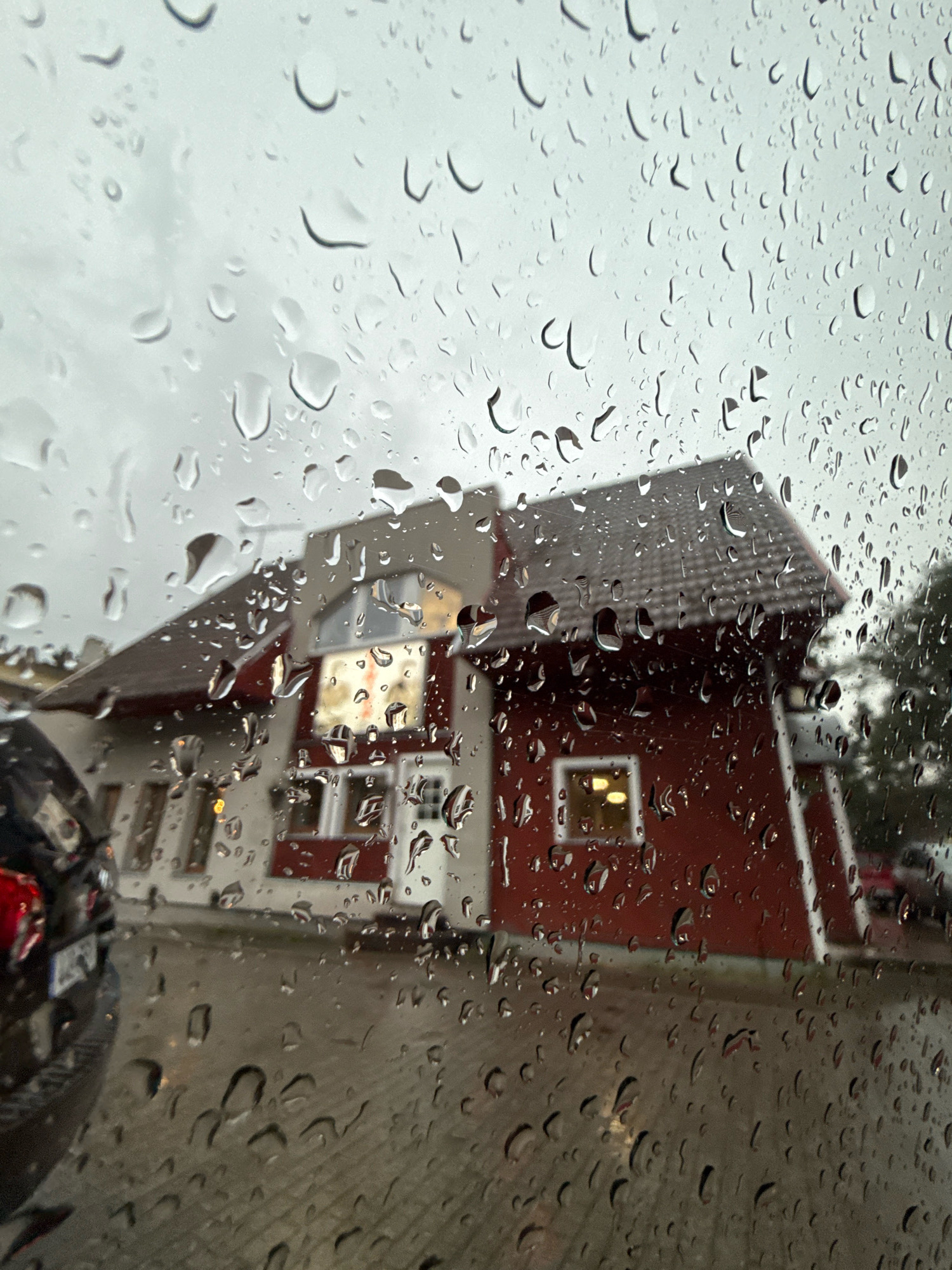 The image depicts a red and white building with a dark roof, possibly a restaurant or shop, seen through a car windshield covered in raindrops. The building has several windows, some of which are illuminated, suggesting it is evening or nighttime. A dark-colored car, seen from the rear, is parked in front of the building. The ground appears to be paved with cobblestones or similar material. The sky is overcast and gray. The scene is likely located in Tallinn, Estonia, as indicated by the prompt. The overall impression is one of a rainy, atmospheric evening in a European city.