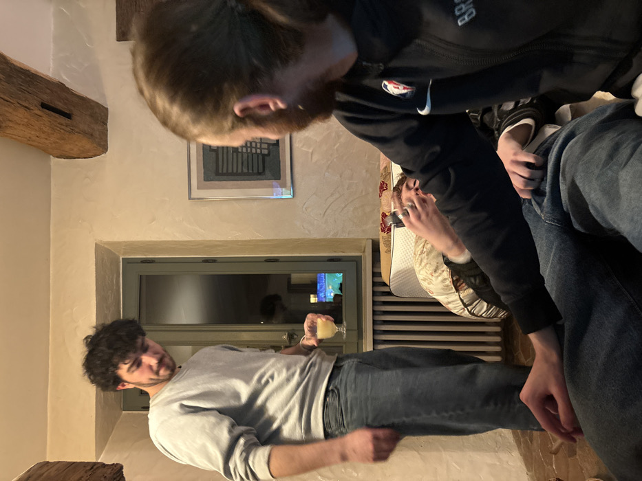 The image depicts an indoor scene in Bû, France, involving three male subjects within a room characterized by rustic architectural elements and artificial lighting.
On the left, a male subject with dark, curly hair and a beard stands, facing partially towards the right foreground. He wears a light grey crew-neck sweatshirt and blue jeans. In his right hand, he holds a stemmed glass containing a pale yellow liquid. His gaze is directed downwards and to the right. Behind this individual, a window with a light green frame reflects interior light sources and displays a distant blue and green light source externally. A white vertical radiator is positioned beneath the window.
In the immediate foreground, a second male subject is seated with his back partially towards the camera. He possesses long brown hair tied back at the nape. He wears a dark zip-up jacket with a white Nike swoosh logo and the word "BROOKLYN" visible on the right chest. His left hand rests on his upper thigh, which is clad in blue denim.
A third male subject is semi-reclined on a patterned sofa or bed positioned between the standing and foreground seated individuals. Only his hands and part of his head are discernible. He holds a silver smartphone with both hands, positioned in front of his face, indicating engagement with the device. He is supported by decorative, patterned cushions.
The room features light-colored, textured walls. A dark, exposed wooden beam is visible in the upper left ceiling area. A framed abstract artwork, composed of dark and grey horizontal and vertical lines, hangs on the wall behind the seated subjects. The floor appears to be constructed of reddish-brown tiles.