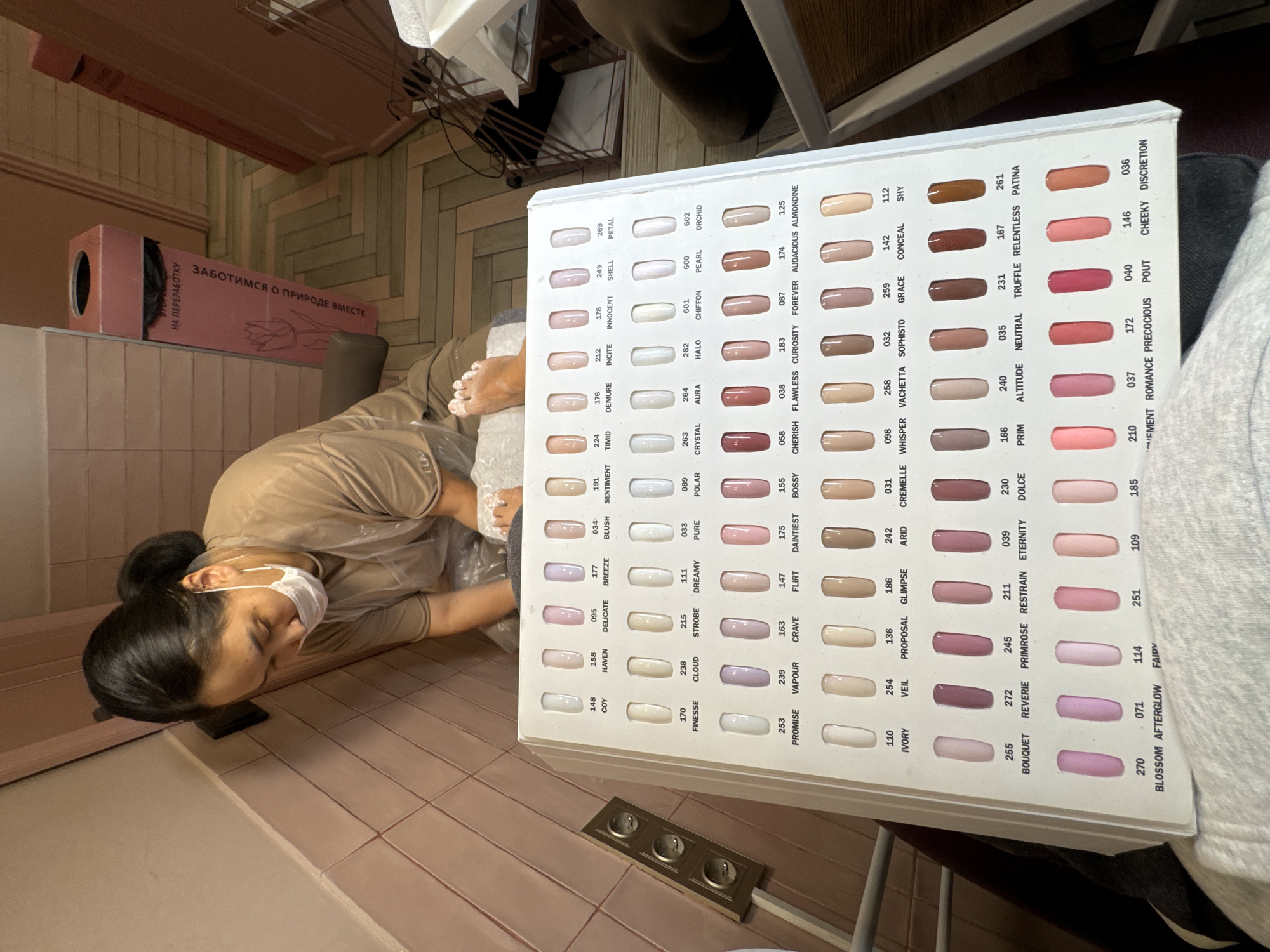 A woman wearing a face mask and a light brown shirt is performing a pedicure on a client. She is sitting in a tiled room with a color palette for nail polish in front of her. The color palette is sitting on a table or chair. There is a box in the background that says "Заботимся о природе вместе" which translates to "Let's take care of nature together" in Russian. The wall has a Russian text - "НА ПЕРЕРАБОТКУ". There is a triple power outlet near the color palette.