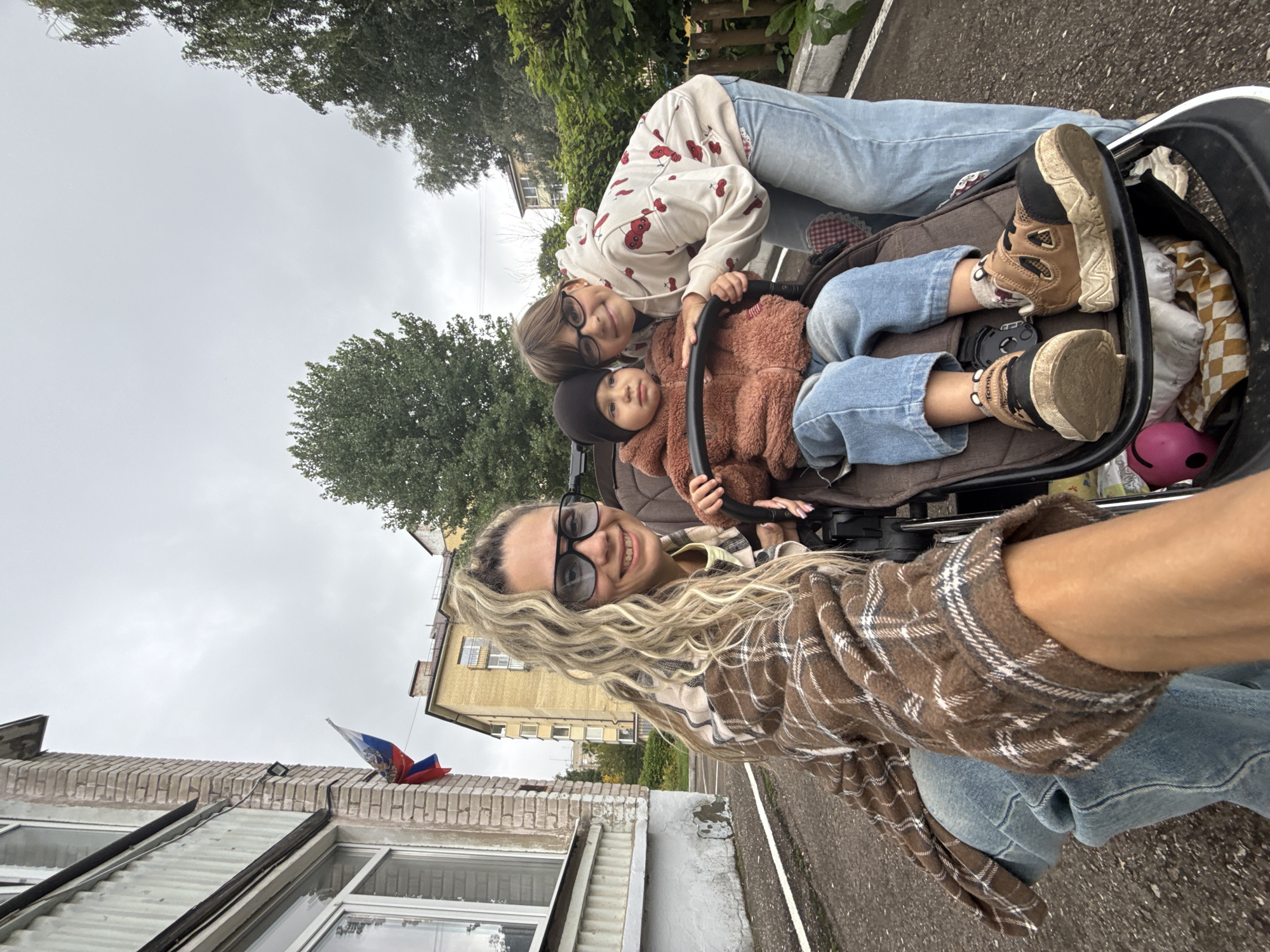 In Rusanovo, Russia, on an overcast day, a woman wearing a brown and white plaid jacket and blue jeans is in the foreground, taking a selfie. She has blonde curly hair and is wearing large black-rimmed glasses and smiling. Beside her, a stroller contains a toddler wearing a brown fuzzy jacket and blue jeans. The toddler is looking ahead with a neutral expression. Behind the stroller, a young girl with dark hair and glasses, wearing a cream-colored hoodie with red cherry prints and blue jeans, leans over and places her arm around the toddler. The background features a building with light yellow walls and windows, and a Russian flag is visible on the building's facade. Trees and greenery line the right side of the scene, and a paved path runs in front of the building. The overall atmosphere is casual and familial.