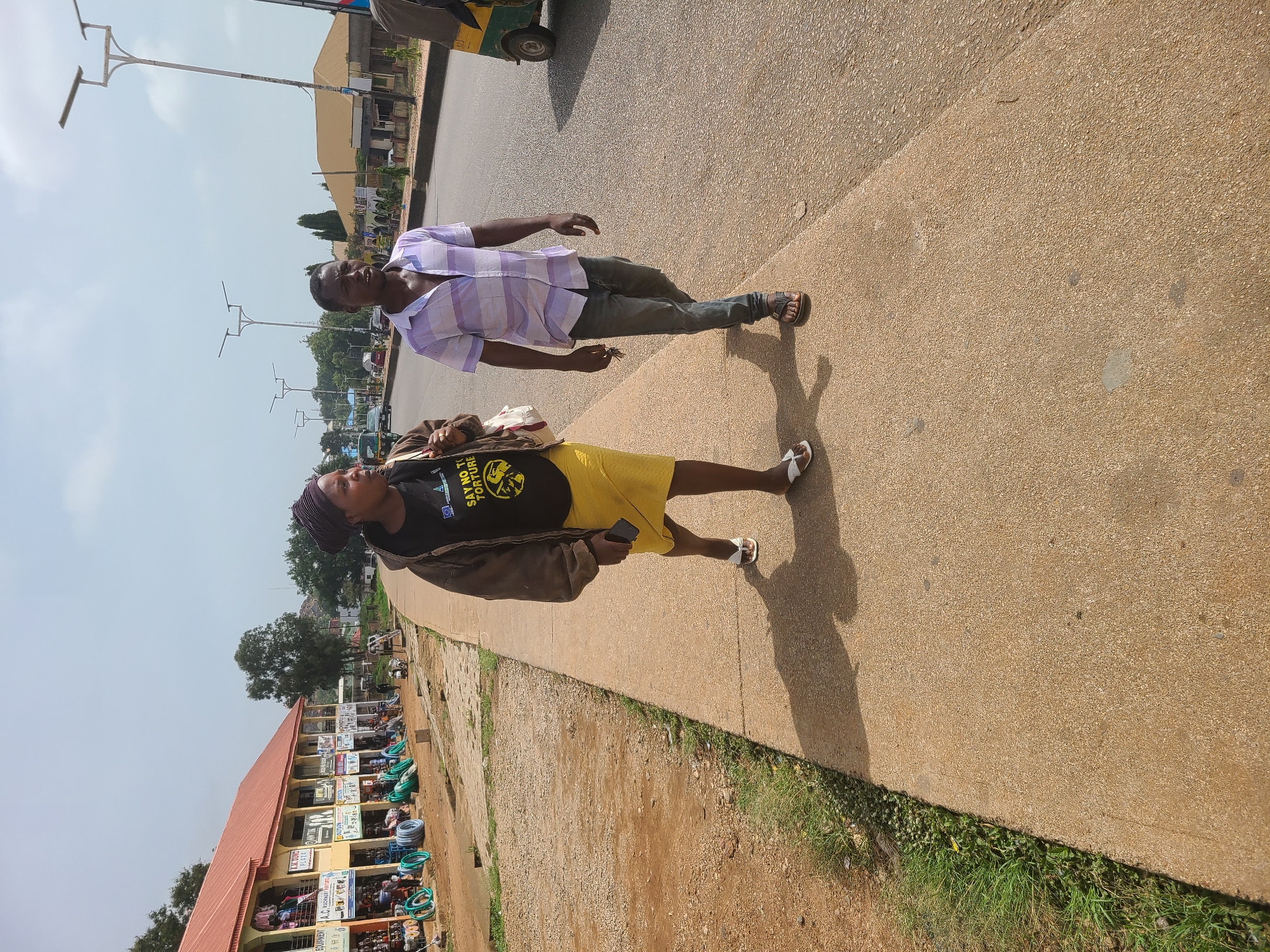 Two individuals, a man and a woman, are walking on a sidewalk next to a road in an urban setting. The man is wearing a light-colored patterned short-sleeved shirt and dark pants, and he is wearing sandals. The woman is wearing a black t-shirt with writing on it, a yellow skirt, a brown jacket and white sandals. She also has a head wrap. They are walking on a wide concrete sidewalk with a curb, next to a road where a vehicle, possibly a tricycle, is partially visible. A low building with numerous storefronts is located along the sidewalk edge. There are a row of light posts behind the man. The weather appears to be sunny. The location appears to be a busy urban area.