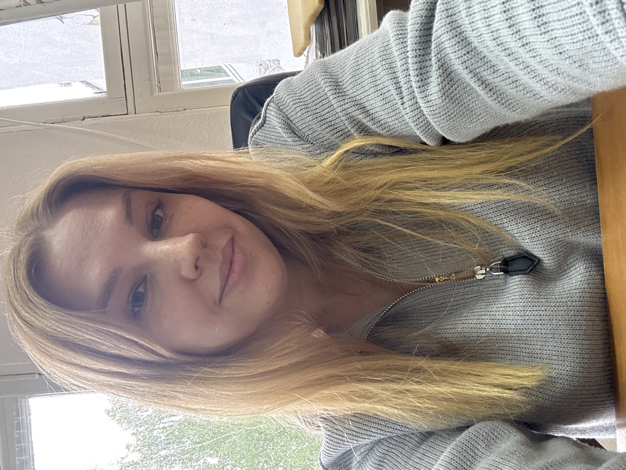 A selfie taken indoors, possibly in an office or home setting, in Rusanovo, Russia. A woman with long, blonde hair is the primary subject. She is wearing a light gray, zip-up sweater. A window is visible in the background, suggesting it is daytime. The background includes glimpses of greenery, indicating an outdoor environment and possible city landscape. A portion of a desk is seen to the right. A black chair is visible in the background.