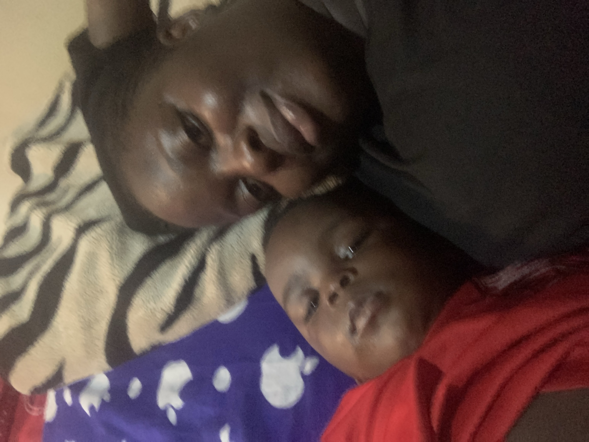 The image shows a close-up, selfie-style shot of two people lying down. A woman with dark skin and dark hair is on the right, looking at the camera with a slight smile. Her right arm is behind her head. To her left, a child with dark skin is also lying down, facing away from the camera, with their eyes open. They are both wearing casual clothing, with the child in a red top and the woman in a black top. The background appears to be a bed with patterned blankets, one with a zebra stripe design and another with a purple background and white cloud-like shapes. The lighting is dim, suggesting an indoor setting, possibly at night or in a dimly lit room. There are no visible landmarks or context to indicate a specific location within a city.