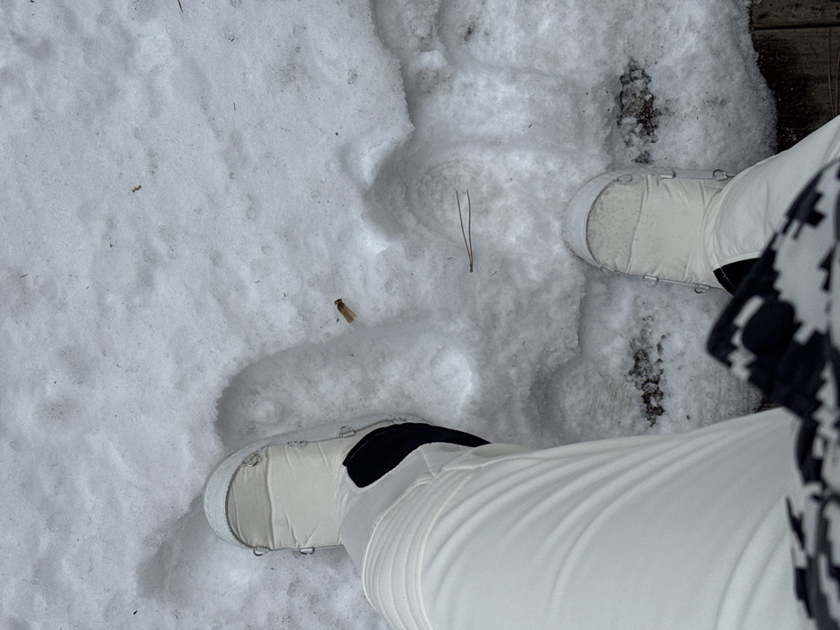 The image depicts a first-person, downward-facing view of a person standing in snow in Courchevel, France. The individual wears white, quilted winter boots with visible metallic eyelets and white, ribbed winter pants. A garment with a black and white geometric pattern is visible at the bottom of the frame, consistent with the wearer's attire. The ground is covered in uneven, possibly disturbed snow, showing footprints or depressions. Visible within the snow are a single brown cigarette butt, located to the right of the left boot, and a lone pine needle positioned horizontally towards the center-right. Darker patches, possibly dirt or melted snow, are present on the ground. A wooden surface, possibly a deck or walkway, is partially visible along the lower and right edges of the frame beneath the snow.