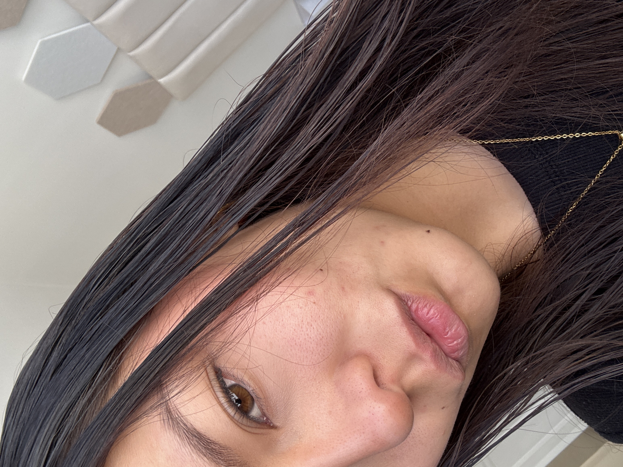 A close-up, angled portrait captures a person, likely female, indoors in San Francisco Ocotlán (Ocotlán), Mexico. The subject's head is tilted, with dark, long hair, appearing somewhat wet or sleek, cascading across the left side of the frame and partially obscuring her face.
Her right eye, a warm brown, is clearly visible and directed towards the camera. The focus is on her facial features, including part of her nose and full, slightly parted lips. Her skin shows natural texture and minor blemishes. A delicate gold-colored chain necklace is visible against a dark, possibly black, garment worn by the subject.
In the blurred background, hexagonal decorative panels in alternating light beige and white tones adorn what appears to be a wall or ceiling. The lighting is soft and even, highlighting the subject's features. There is no discernible activity other than posing for the portrait. No text is visible in the image.