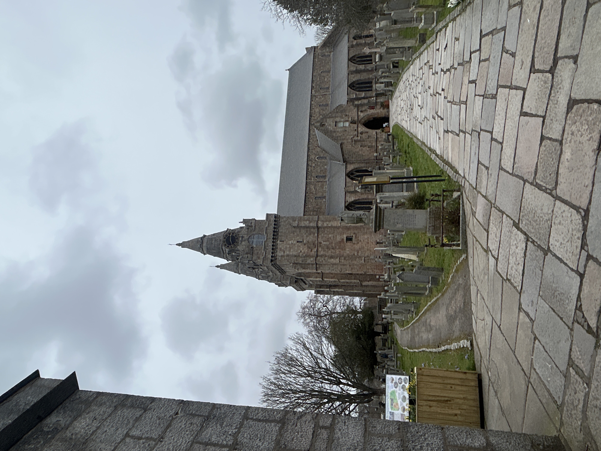 The image depicts St Machar's Cathedral in Aberdeen, Scotland. A stone pathway leads to the cathedral, lined with gravestones of the adjacent cemetery. The cathedral is made of grey stone, featuring a tall tower with a spire. The sky is overcast. A stone wall is in the lower left of the image. A sign stands near the pathway. Bare trees dot the scene.
