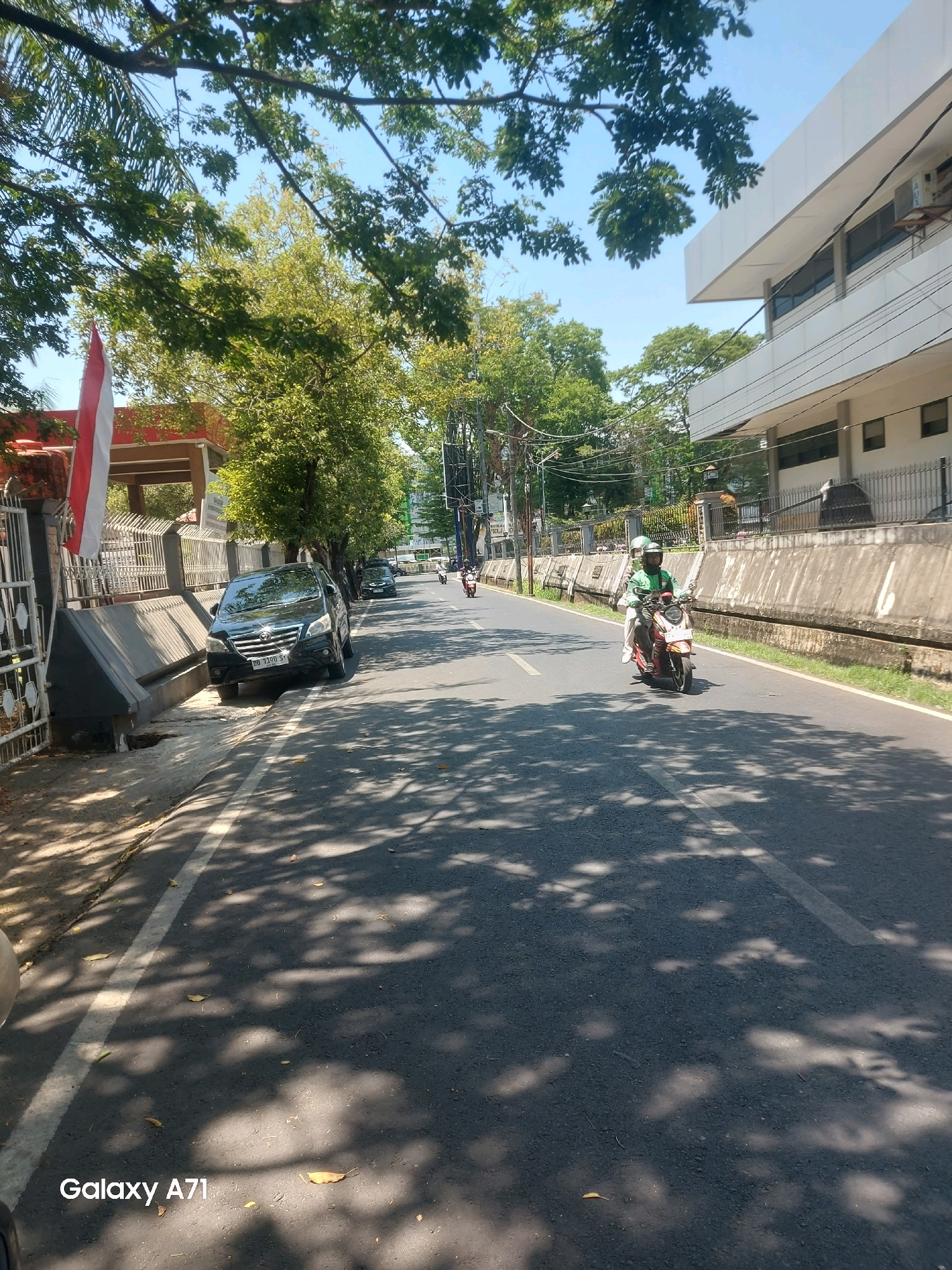 The image depicts a street scene in Makassar, Indonesia, on a sunny day. The foreground is dominated by an asphalt road with dappled sunlight filtering through the leaves of trees lining the street. A white line marks the edge of the road. In the lower left corner, the watermark "Galaxy A71" is visible.
A black car, identified by the license plate "DD 1300 SY", is parked on the left side of the road, partially on the sidewalk. Next to the car, a metal fence and a red and white Indonesian flag are visible, suggesting a government building or official establishment.
Further down the road, a motorcycle with two occupants is traveling away from the camera. The driver, wearing a green shirt and a helmet, is visible. The passenger is also wearing a helmet. Other vehicles, including a car and another motorcycle, are also seen in the distance on the road.
The right side of the street features a concrete wall, a row of grass, and a modern building with multiple stories and large windows. Trees are scattered throughout the scene, providing shade and adding to the natural elements of the urban environment. The sky is clear and blue, indicating fair weather. The overall impression is of a typical day in a bustling urban area of Makassar.