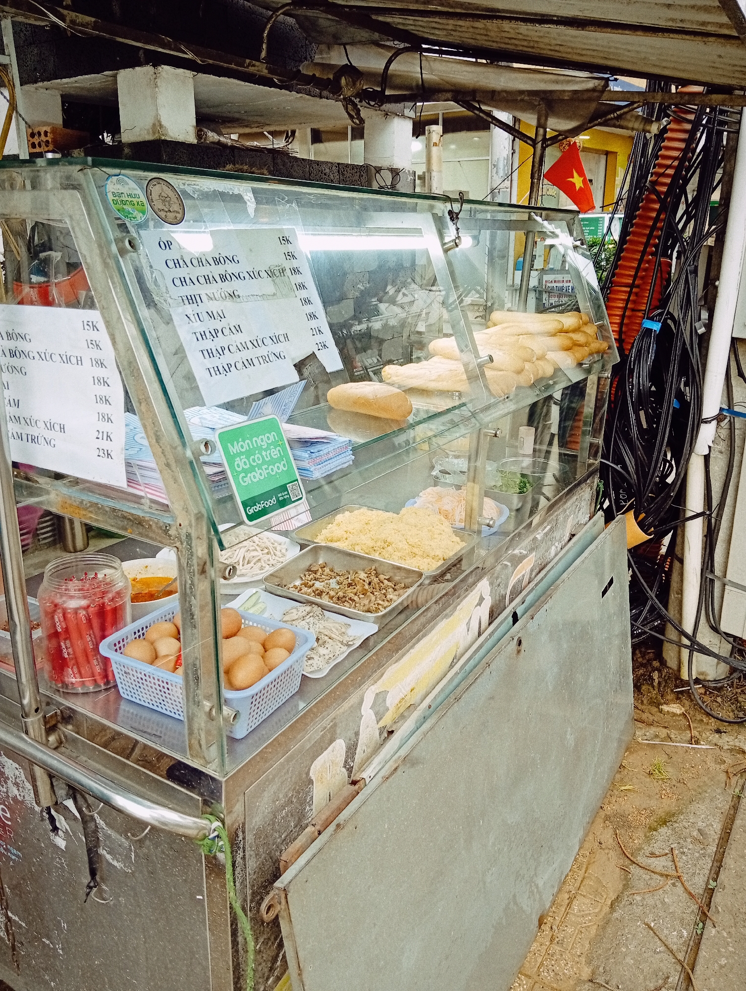 This eye-level shot depicts a vibrant street food stall, likely selling *bánh mì*, set up outdoors in Da Lat, Vietnam. The central subject is a glass-fronted display case atop a stainless steel cart, showcasing various ingredients and fresh baguettes. Inside the case, rows of golden-brown baguettes are neatly arranged. Below, multiple metal trays hold prepared fillings such as shredded pork floss, minced meats, bean sprouts, and what appears to be diced cucumber and herbs. A blue basket filled with brown eggs and a jar of red sausages are visible on the left side of the counter.
A laminated menu is prominently displayed inside the glass case, listing items and their prices in Vietnamese Dong (indicated by "K"). Visible options include "CHA CHÀ BÔNG" (pork floss *bánh mì*) for 15K, "THỊT NƯỚNG" (grilled pork) for 18K, "XÍU MẠI" (pork meatballs) for 18K, and "THẬP CẨM" (mixed/combination) for 18K, with variations like "THẬP CẨM XÚC XÍCH" (combination with sausage) for 21K and "THẬP CẨM TRỨNG" (combination with egg) for 23K. A "Món ngon đã có trên GrabFood" (Delicious dishes are now on GrabFood) sticker with a QR code indicates the stall offers delivery services.
To the upper right, a small red Vietnamese flag with a yellow star adds a distinct local touch. The background is characterized by a dense tangle of black electrical wires and cables, typical of urban infrastructure in Vietnam, running alongside a white pipe. The ground visible next to the cart appears to be concrete with some dirt and small weeds, suggesting a well-used public space. No people are visible, but the neatly arranged food and clear menu suggest the stall is open for business.