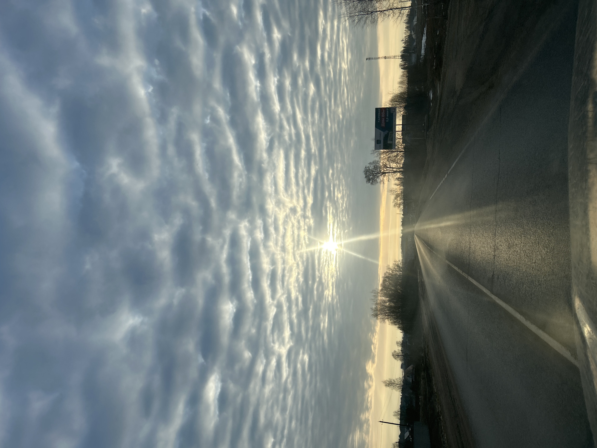 The image shows a view from a vehicle driving on a road in Фоки, Russia. The road stretches towards the horizon with a single white line marking its edge. On the right side of the road, there is a roadside with trees and sparse vegetation. In the distance, a billboard with Russian text is visible. The sky is filled with a pattern of altocumulus clouds, backlit by the sun, which is low on the horizon, casting a glare onto the road. The overall impression is that of a rural or suburban landscape during a sunny day with interesting cloud formations.