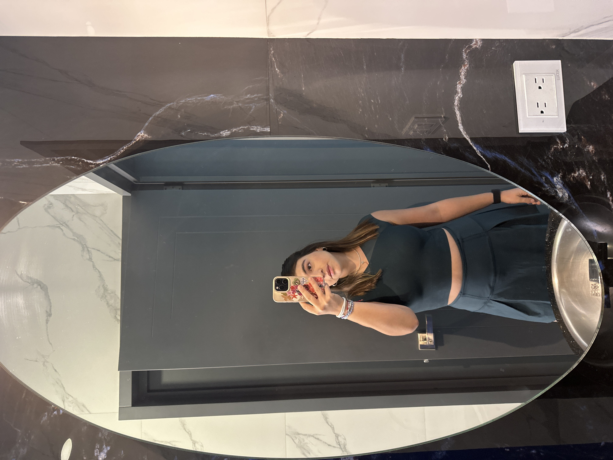 A woman is taking a selfie in a bathroom mirror. The mirror is oval-shaped and reflects her from the waist up. She is wearing a dark green two-piece outfit and holding a smartphone with a floral case. The bathroom has dark marble countertops, white and gray marble walls, and a dark gray door. A white electrical outlet is visible on the counter. The location is presumably in Puebla, Mexico, but there are no specific details to confirm this.