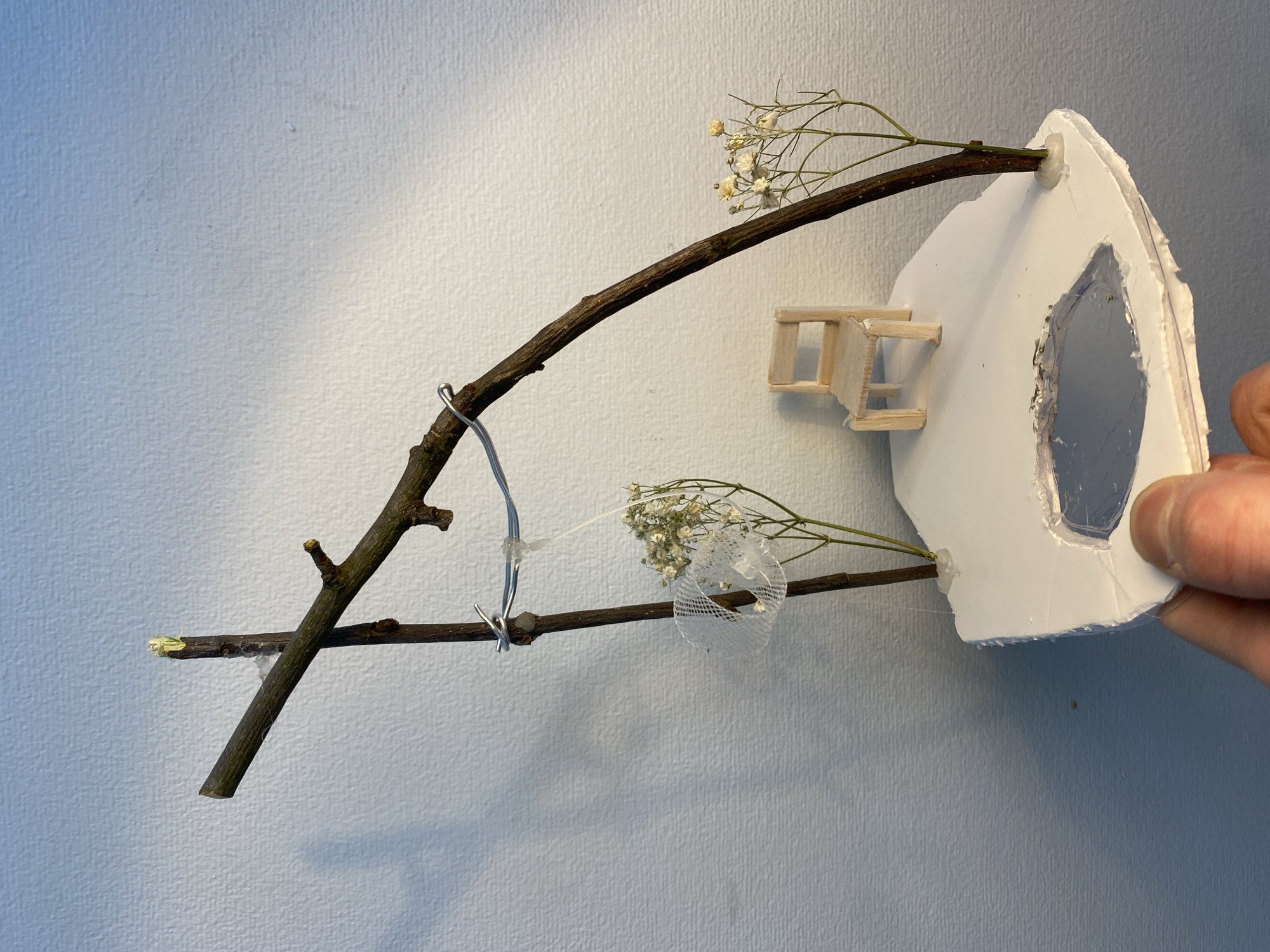 A constructed, whimsical diorama is held in a person's hand against a pale blue-grey wall. The diorama features two brown sticks forming an angled "A" shape. The top stick is adorned with tiny white flowers and is attached to a white, crudely-shaped, house-like structure, possibly made of foam or plastic. The lower stick also has similar white flowers and a small piece of white netting attached to it. A wire is looped around the two sticks, connecting them. A miniature wooden ladder is affixed to the side of the white structure. The white structure has an irregular hole filled with a clear material, possibly plastic or resin, that serves as a window. The background is a smooth, solid-colored wall. There is nothing to suggest the location in the city of Moscow, Russia.