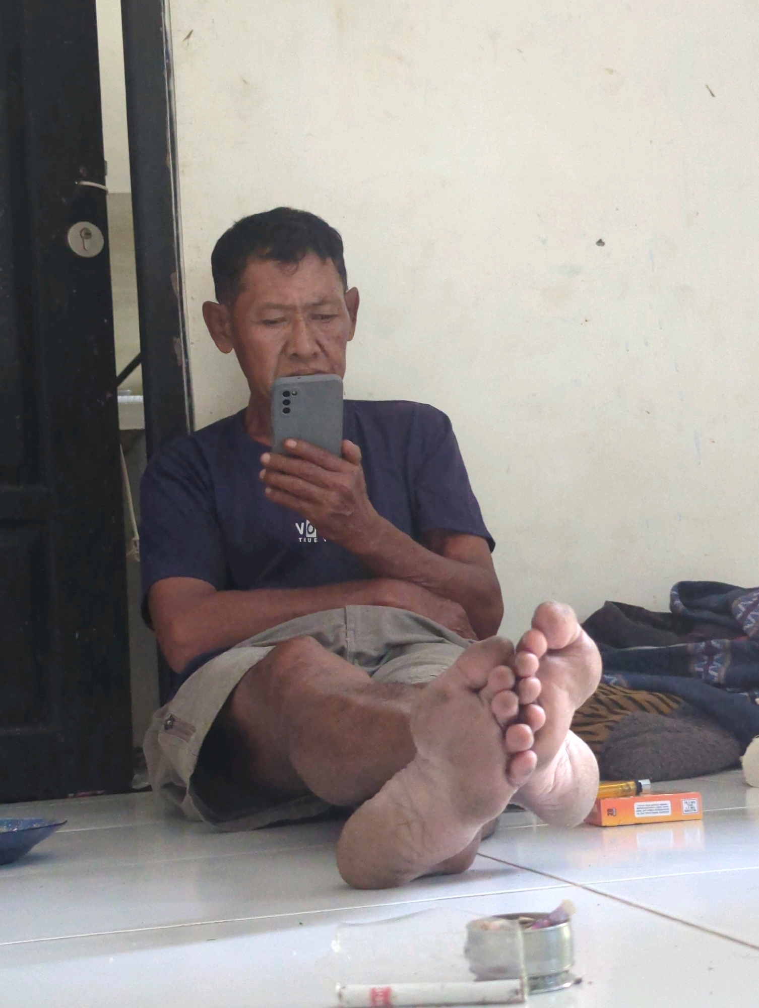 In Batu Belaman, Indonesia, a man is sitting on the floor near a doorway, looking at his smartphone. He is wearing a dark blue t-shirt and light-colored shorts. His feet are bare and prominently displayed in the foreground. In the background, a wall is visible, along with a folded blanket or rug and a pack of cigarettes. There is also an ashtray with cigarette butts and another cigarette lying on the floor. The scene appears to be an outdoor or semi-outdoor area, possibly a porch or patio.