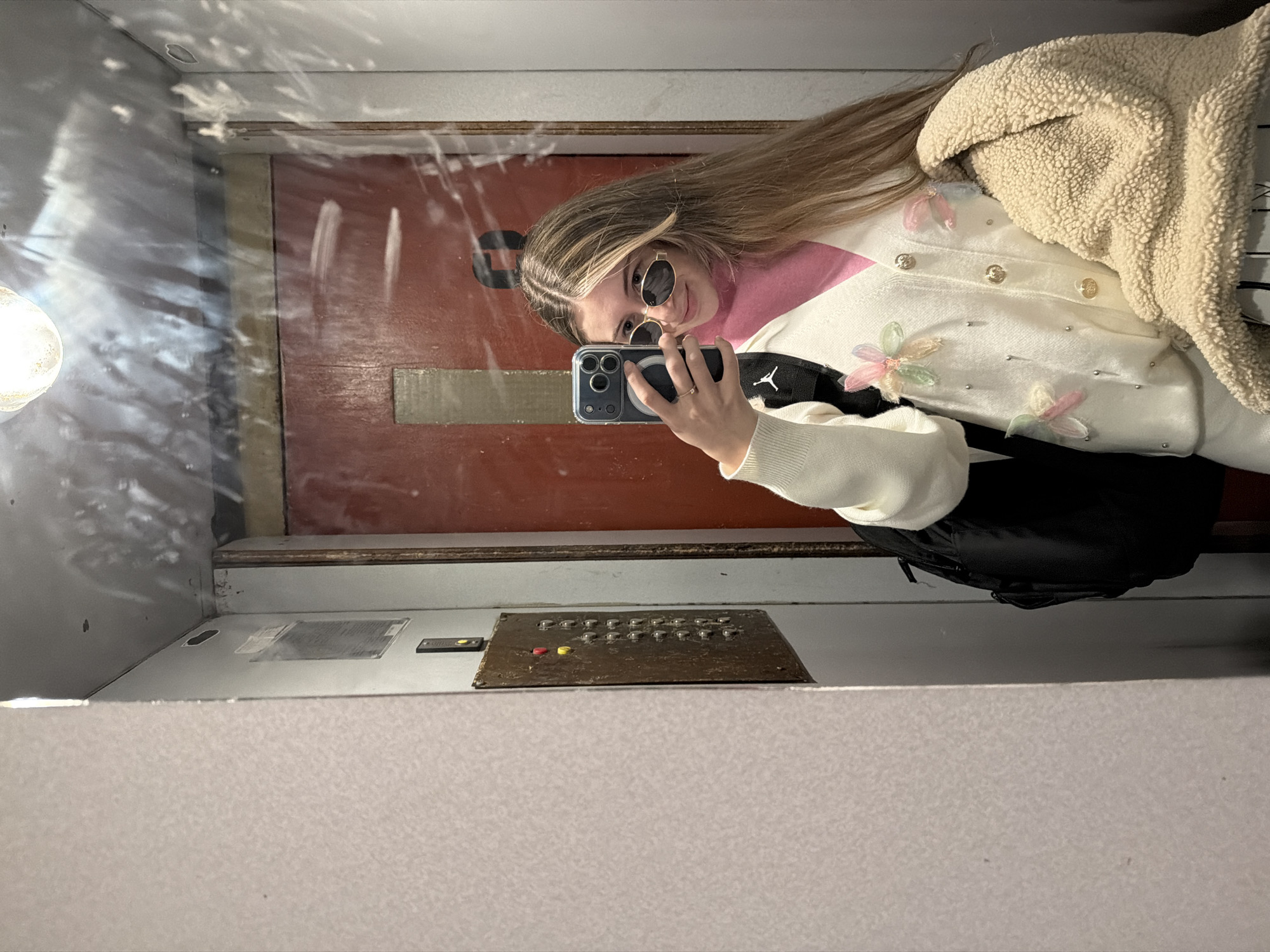 A woman with long blond hair is taking a selfie in an elevator. She is wearing round sunglasses, a pink turtleneck, a white cardigan with flower embellishments, and a beige sherpa jacket. She is holding a blue iPhone in her left hand. The elevator appears old, with a dark red door and an old-fashioned control panel with multiple buttons. The interior of the elevator is mostly gray, with a light fixture visible at the top. This could be in a public building in Plovdiv, Bulgaria, considering the architecture and context.